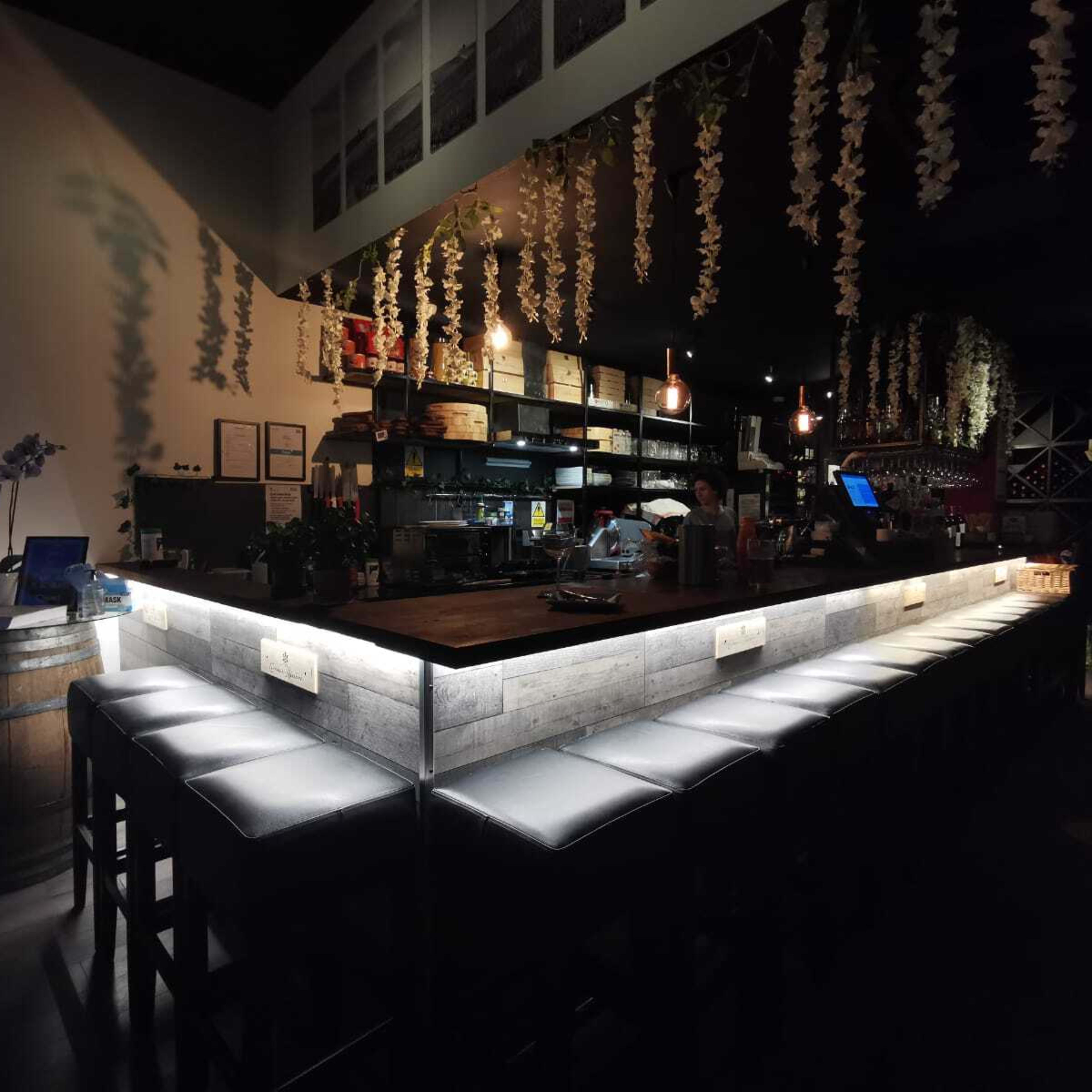 A dimly lit bar features a long, illuminated counter with high stools and shelves lined with various bottles.