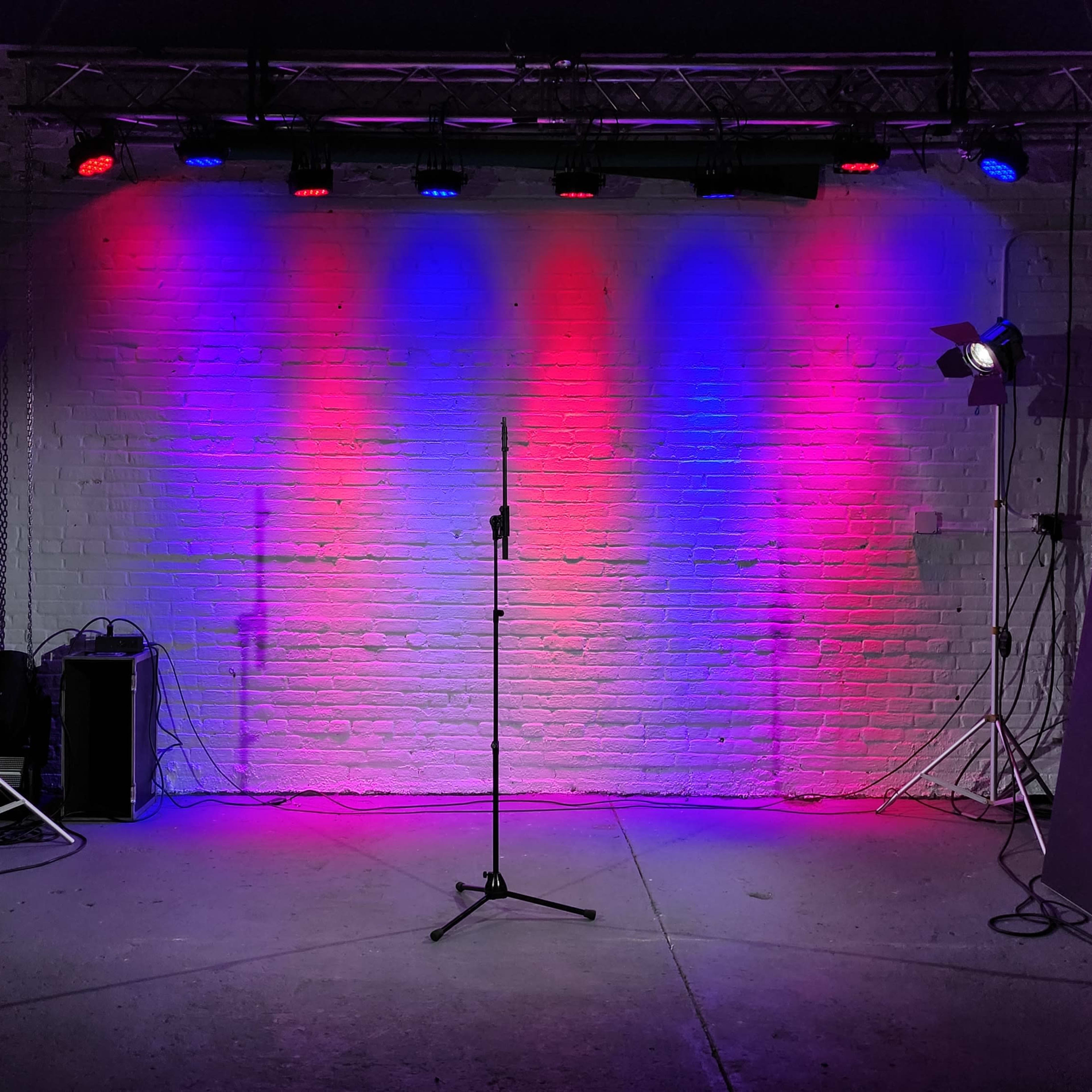A performance stage with colored lights illuminating a microphone stand in front of a lit white brick wall.
