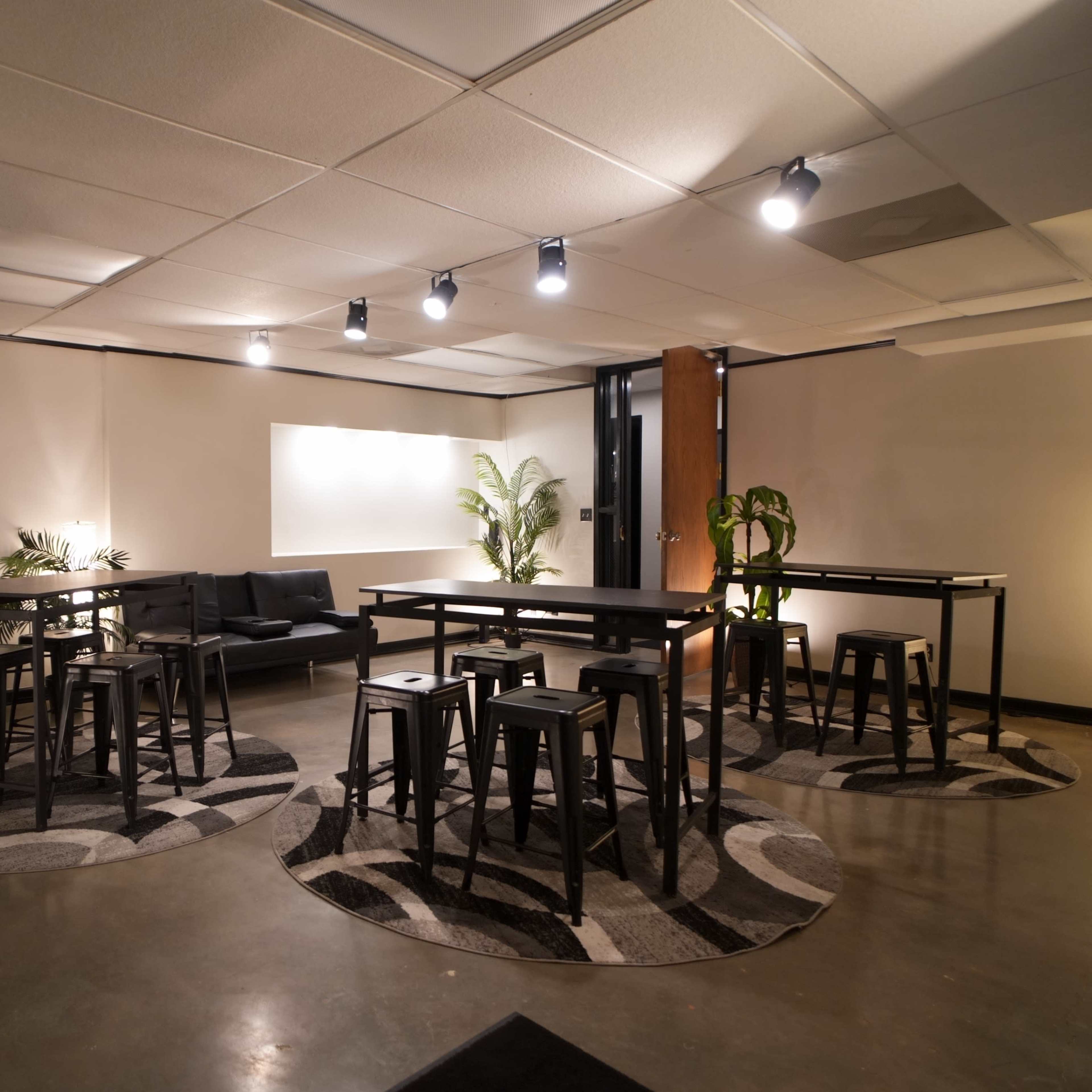 A modern lounge area featuring several high tables and stools arranged on patterned rugs, illuminated by ceiling lights.
