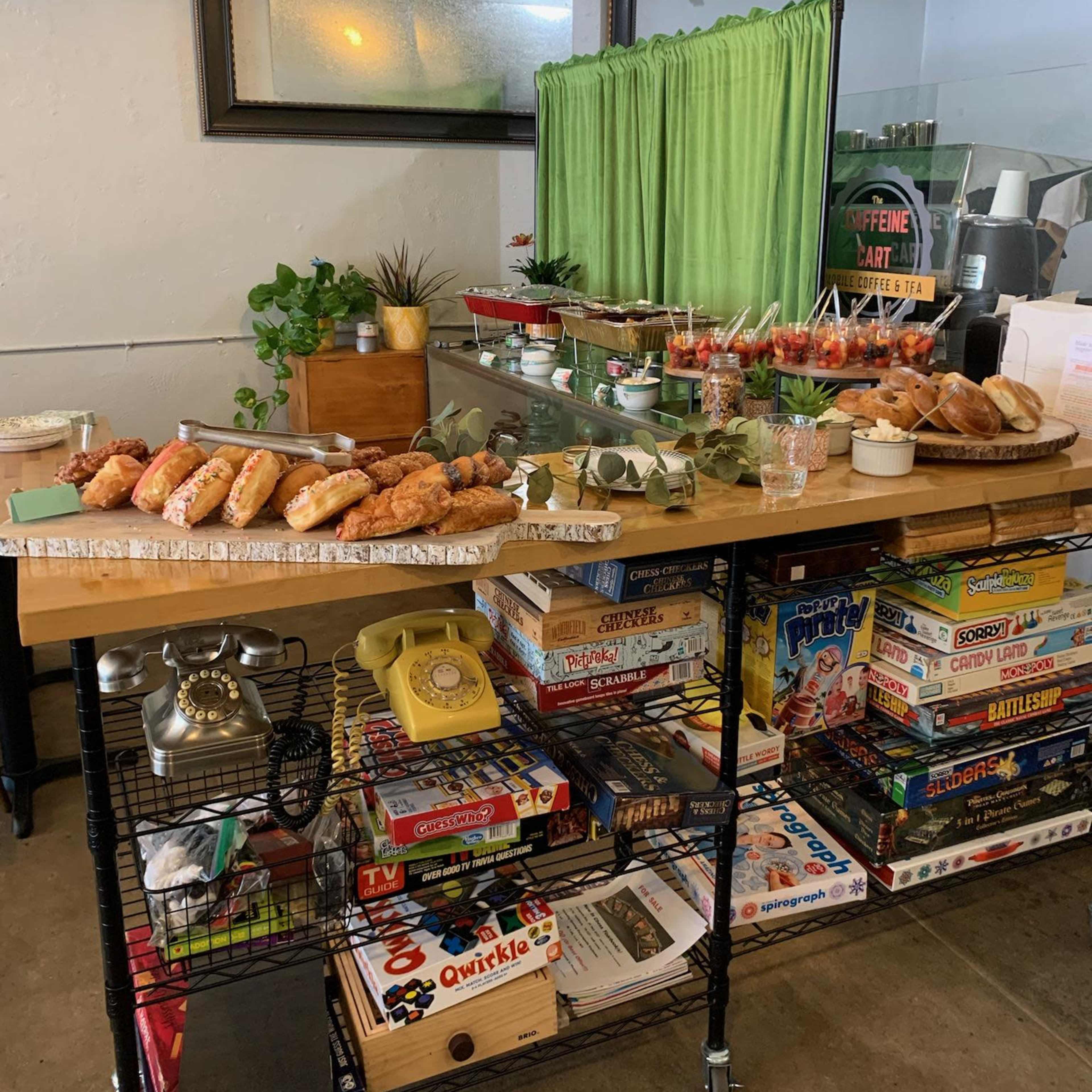 A table loaded with various baked goods and snacks is set up next to a green curtain and a shelf of board games.