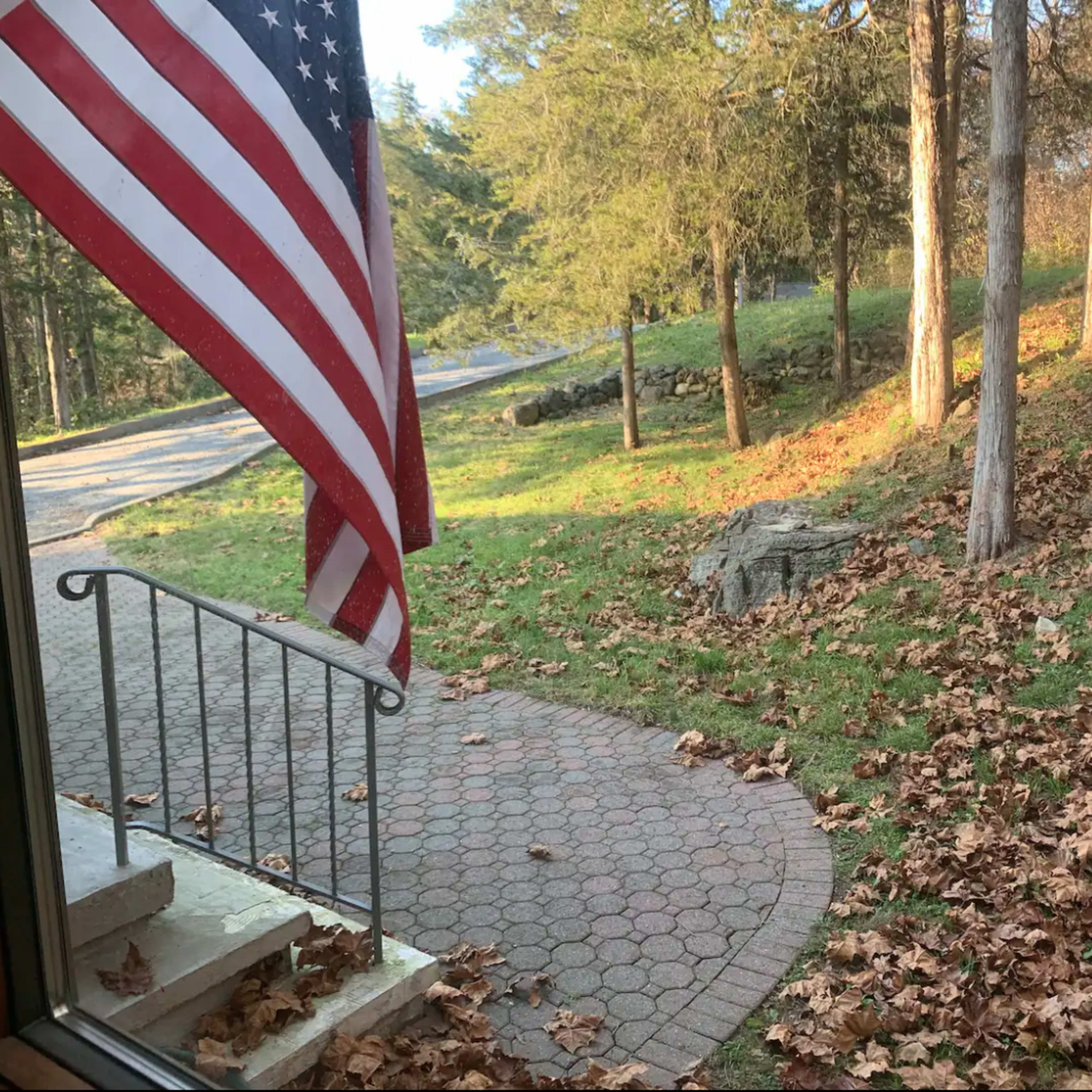 An American flag waves beside a stone pathway covered with autumn leaves, leading to a tree-line view and a road.