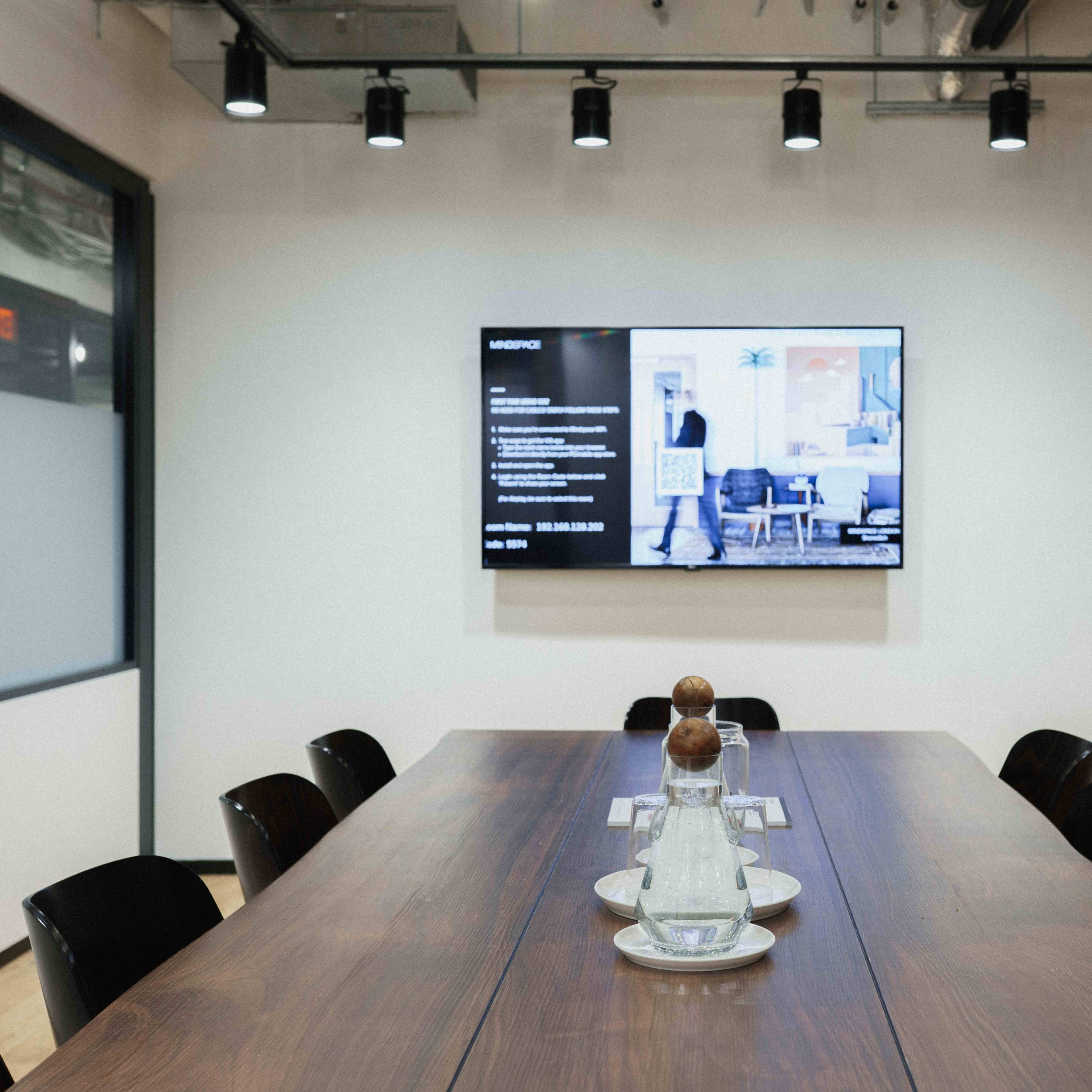A conference room features a long wooden table, a large wall-mounted screen displaying information, and black chairs arranged around the table.