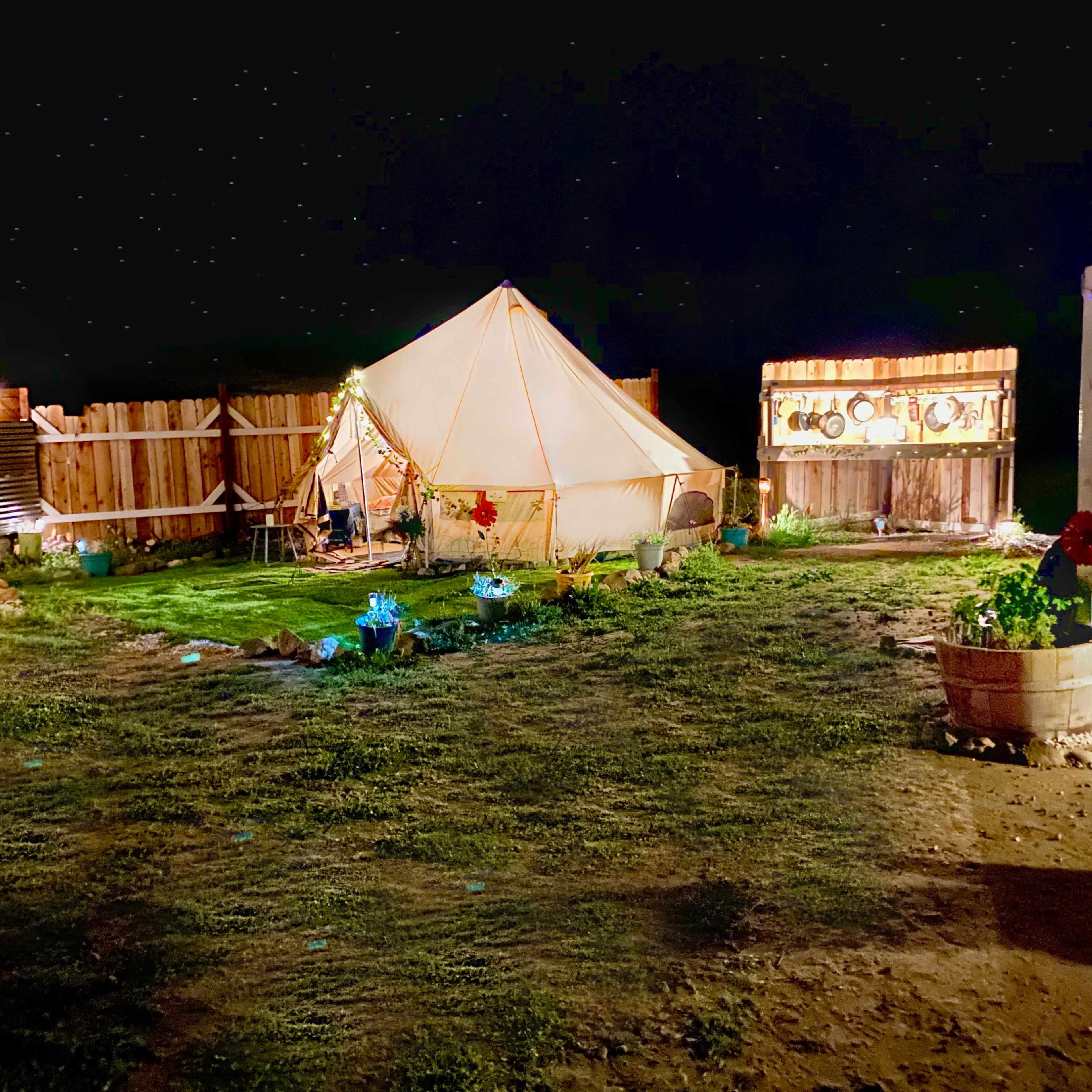 A large canvas tent is set up in a grassy area surrounded by wooden fencing, with decorative lights and potted plants illuminating the scene at night.