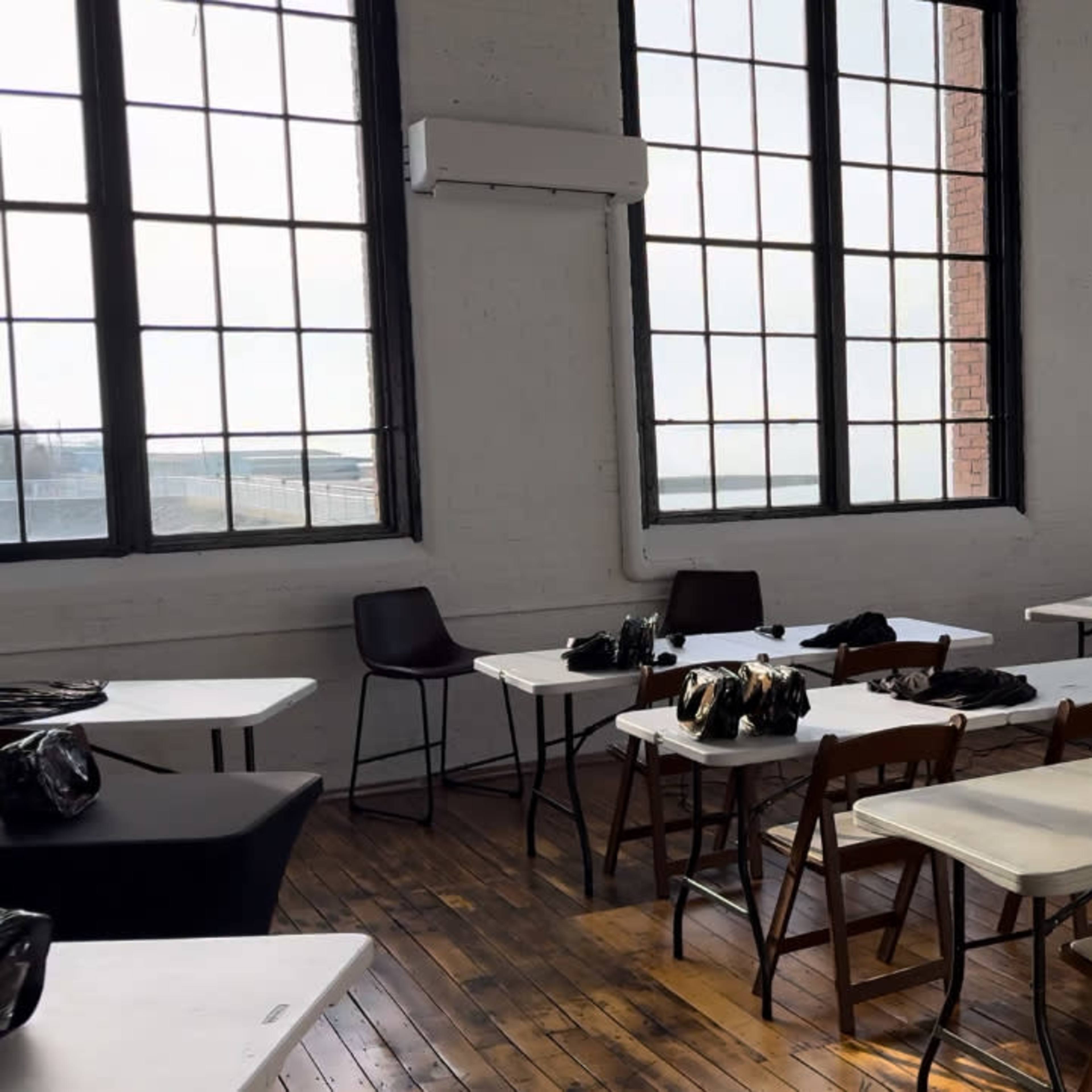 A room with large windows features several tables and chairs, each equipped with bags and cameras, indicating a setup for a workshop or event.