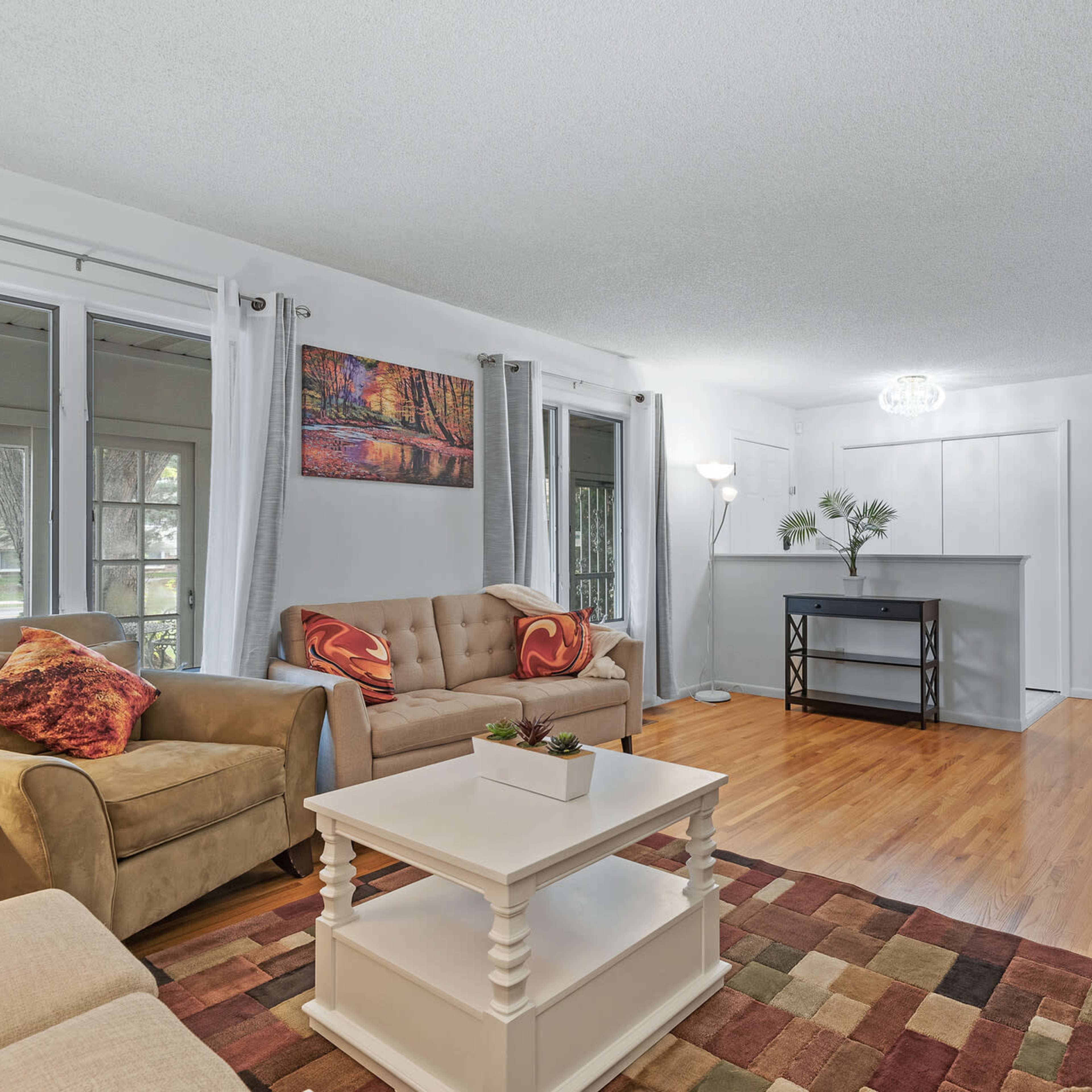 A bright living room with two sofas, a coffee table, and a bar area, featuring wooden floors and large windows.