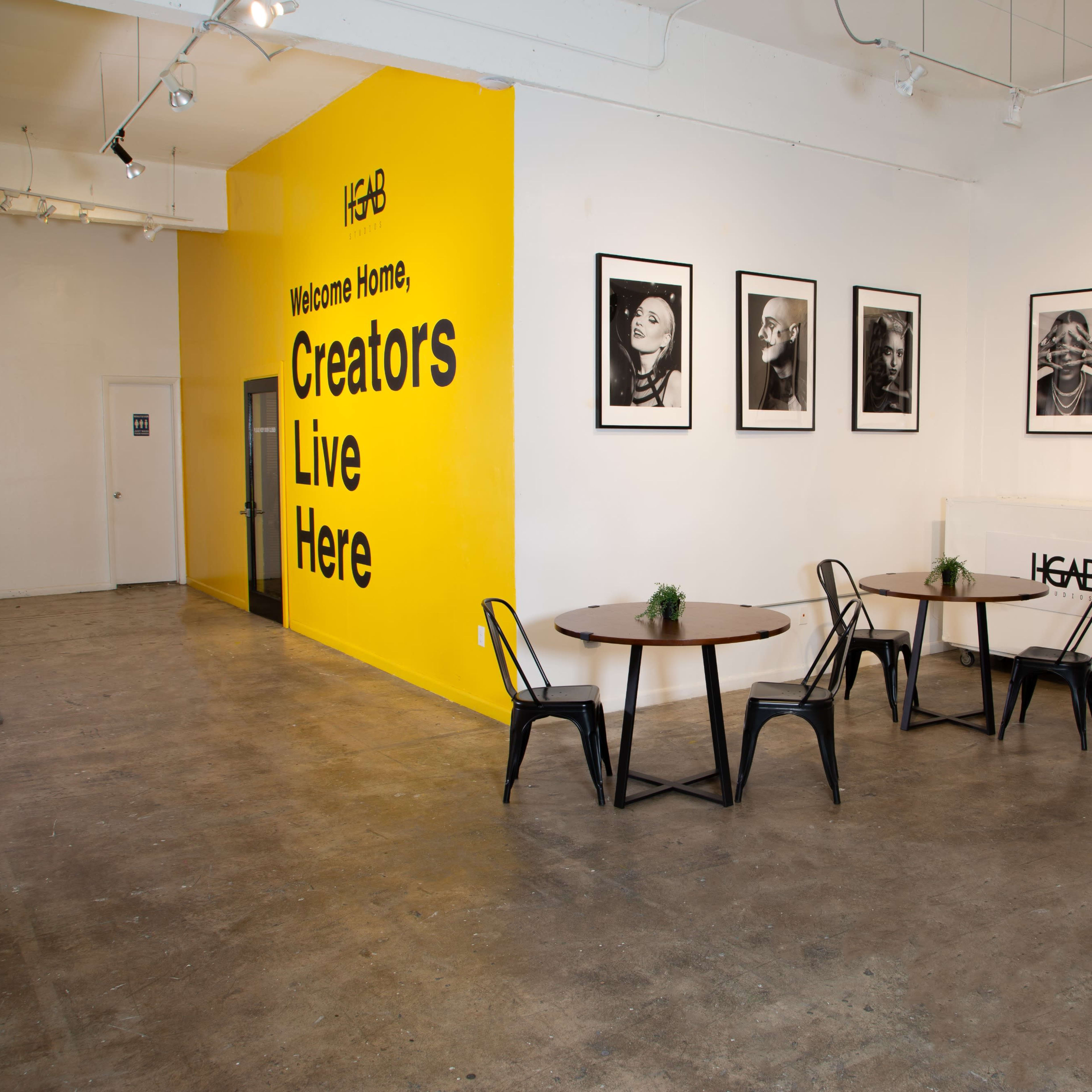 A bright yellow wall with black framed portraits, two round tables with chairs, and an entryway in a spacious interior.