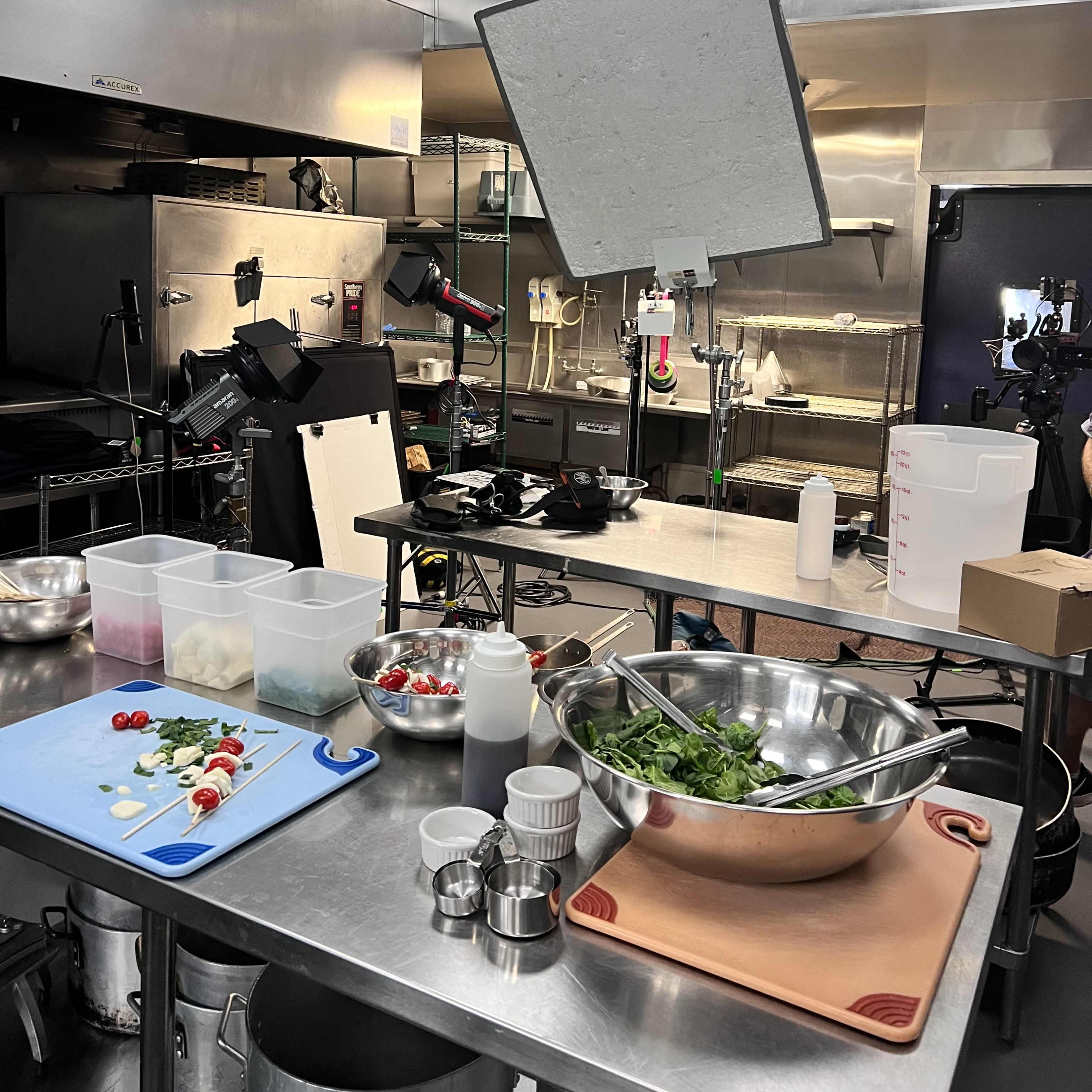 A commercial kitchen setup with cooking equipment, a variety of ingredients on a countertop, and lighting equipment arranged for a photoshoot.
