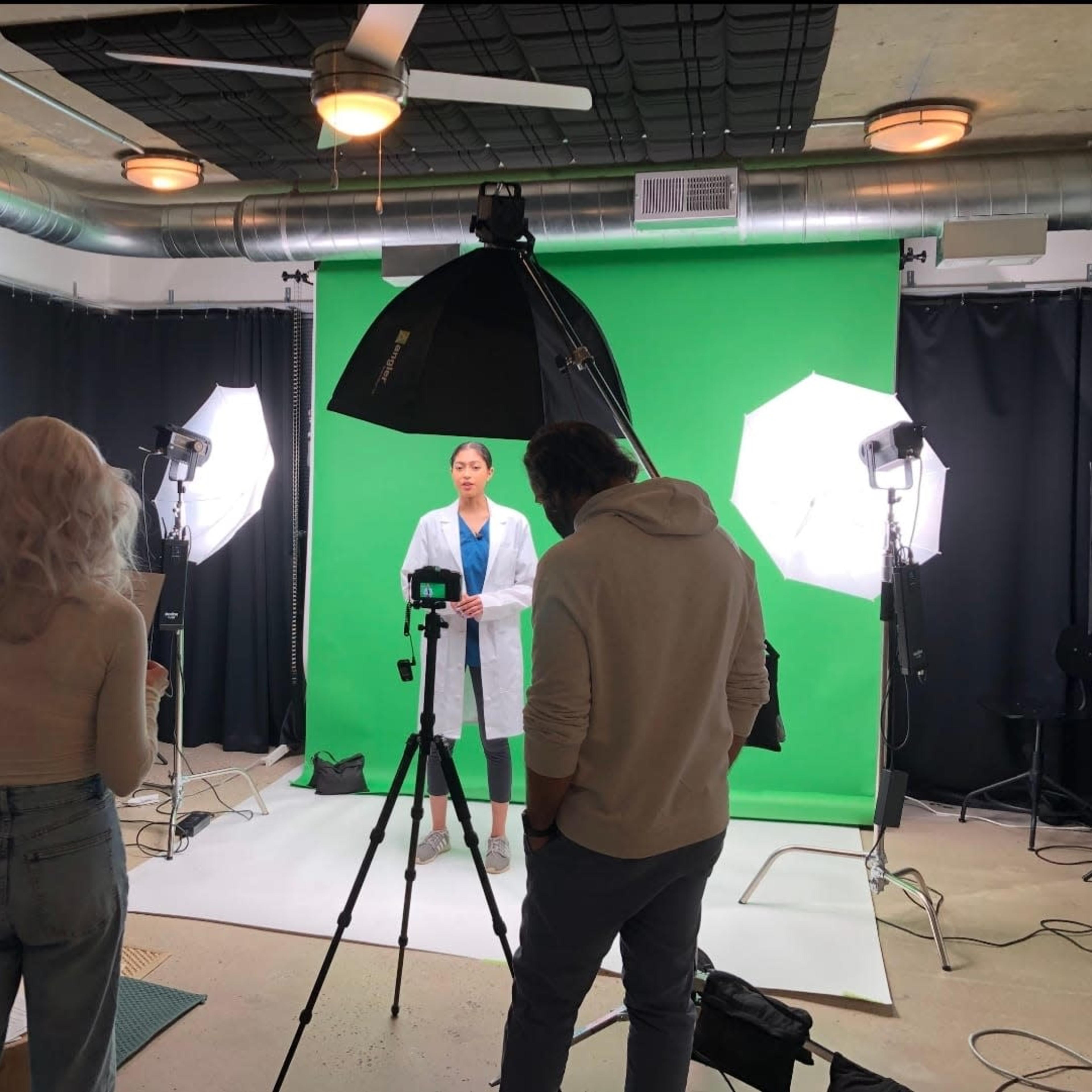 A woman dressed in a lab coat stands in front of a green screen while two individuals operate a camera and lighting equipment in a studio setting.