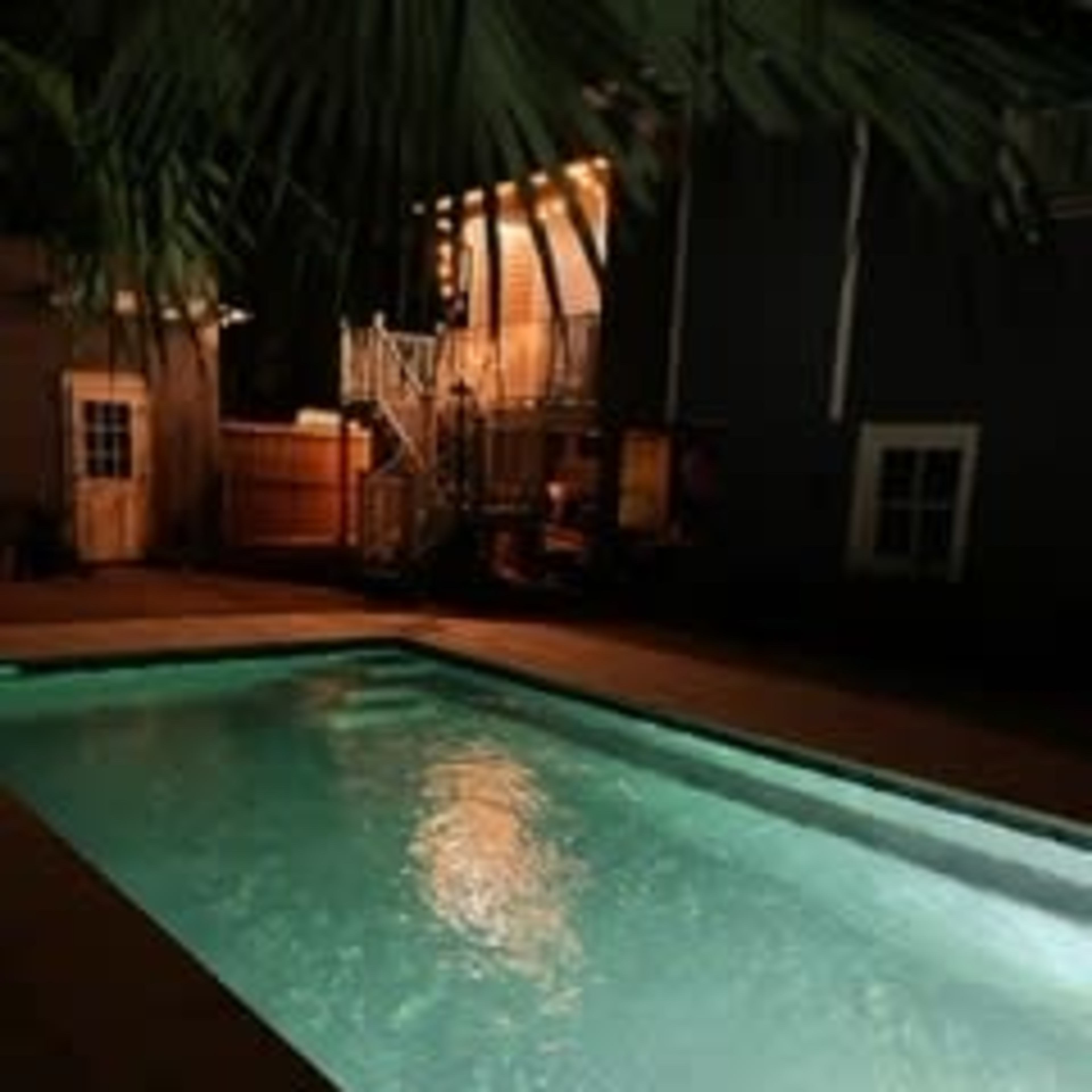 A well-lit swimming pool with clear water is surrounded by palm trees and a house illuminated by warm lights at night.