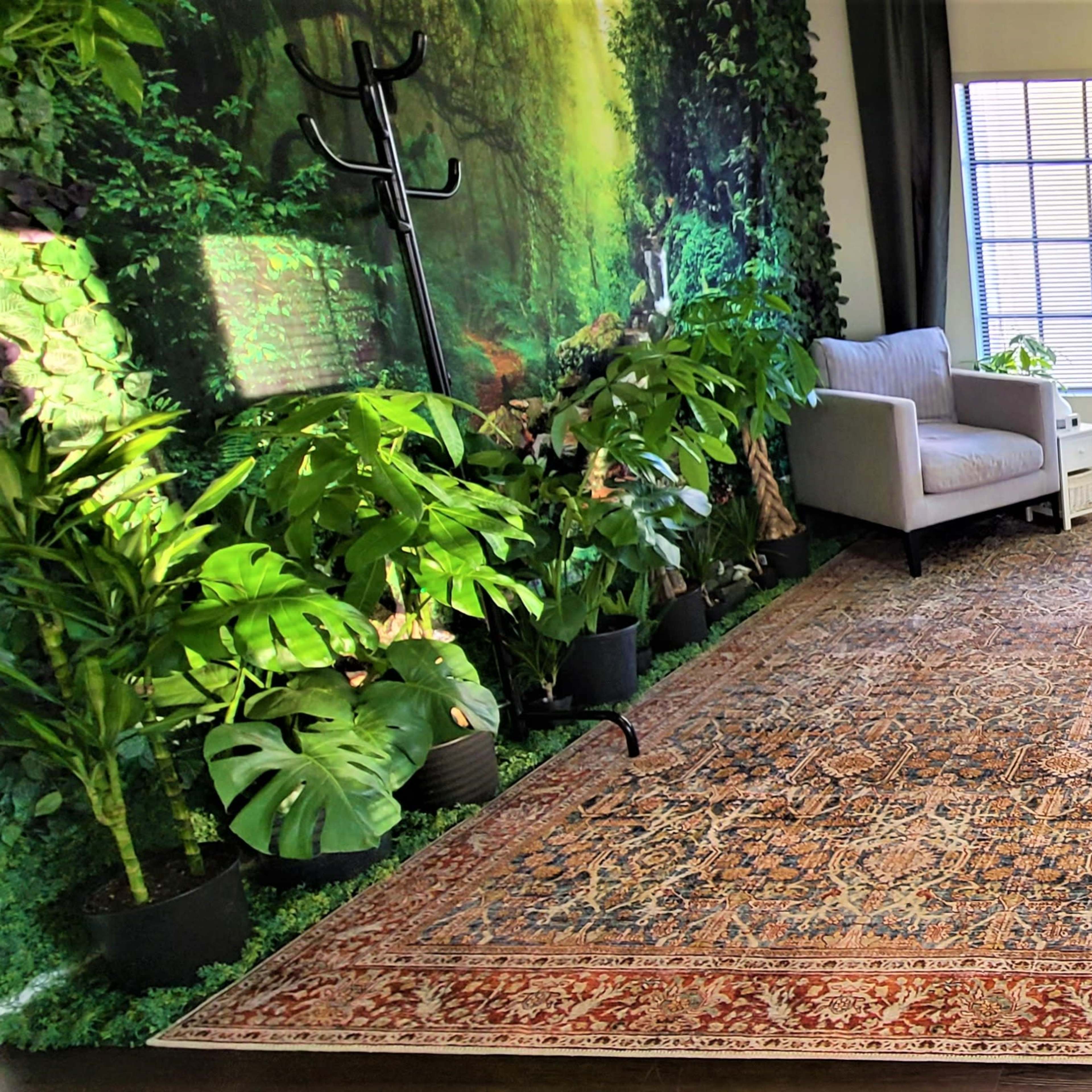 A room featuring a patterned rug, a gray armchair, a desk with a chair, and a wall adorned with greenery and windows.