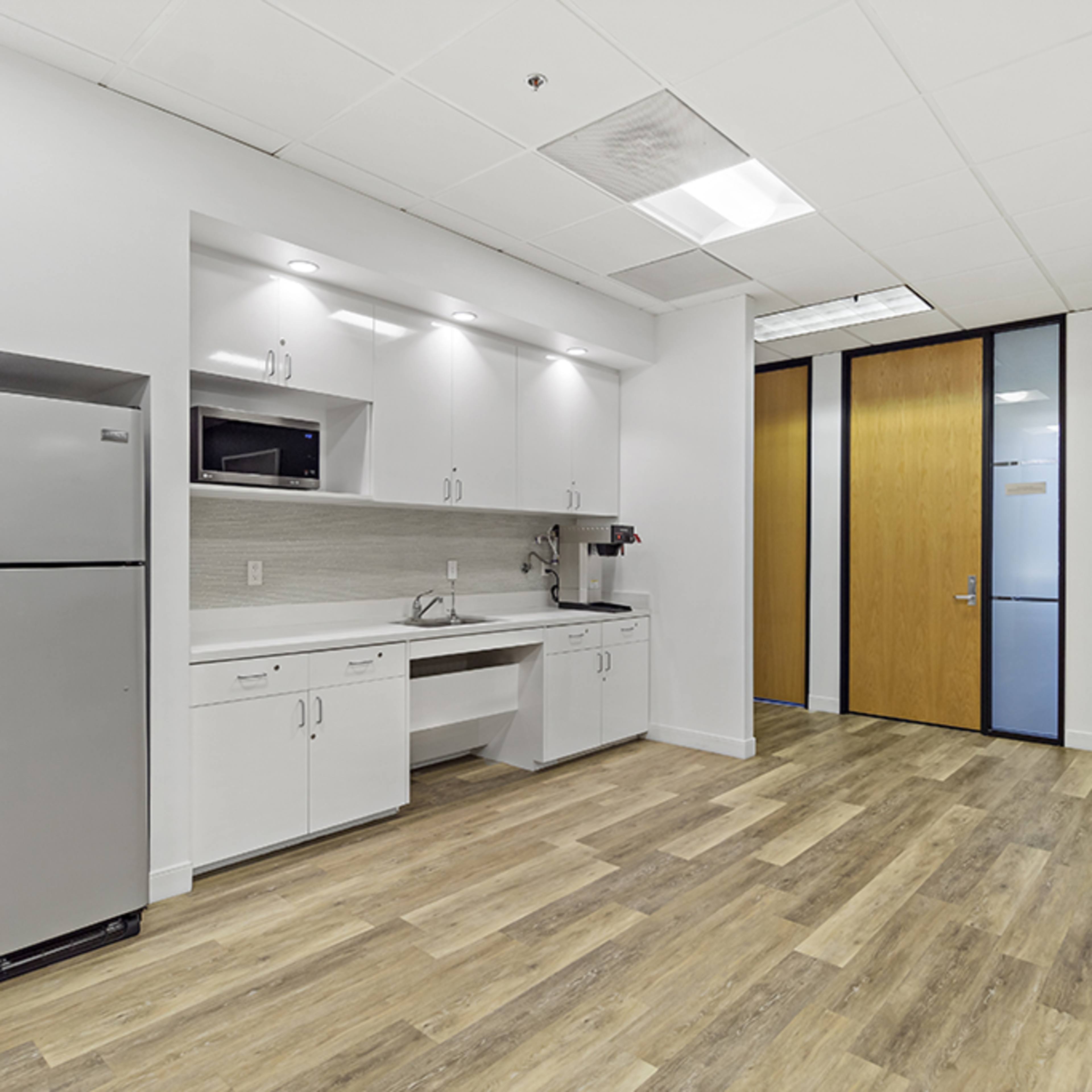 A modern kitchenette with a refrigerator, sink, and microwave is situated in a room adjacent to a glass-paneled office door.