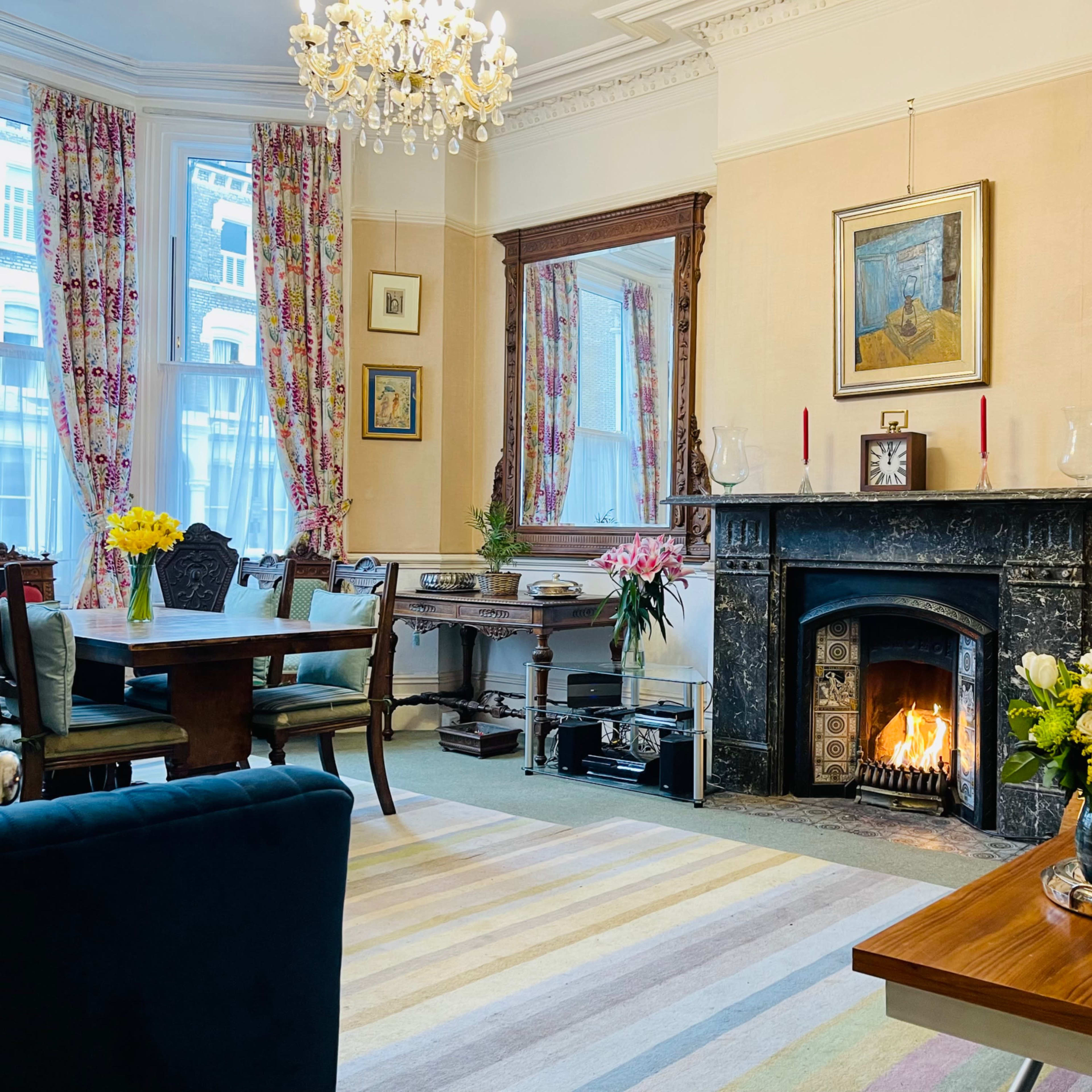 A cozy living room features a fireplace, a dining area with floral decorations, and a large window adorned with patterned curtains.