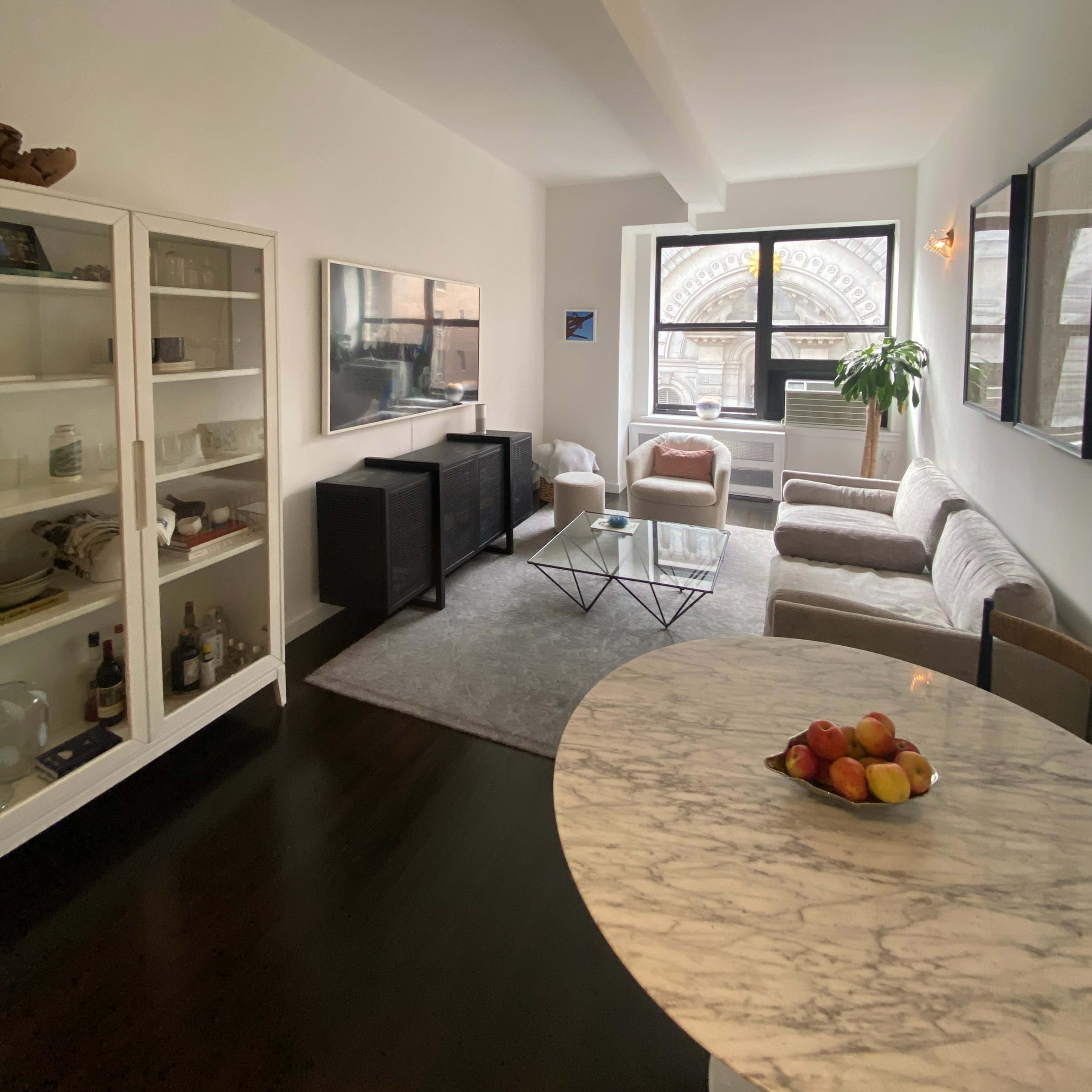 A modern living room featuring a sofa, coffee table, television, and a nearby dining area with a marble tabletop and a bowl of fruit.