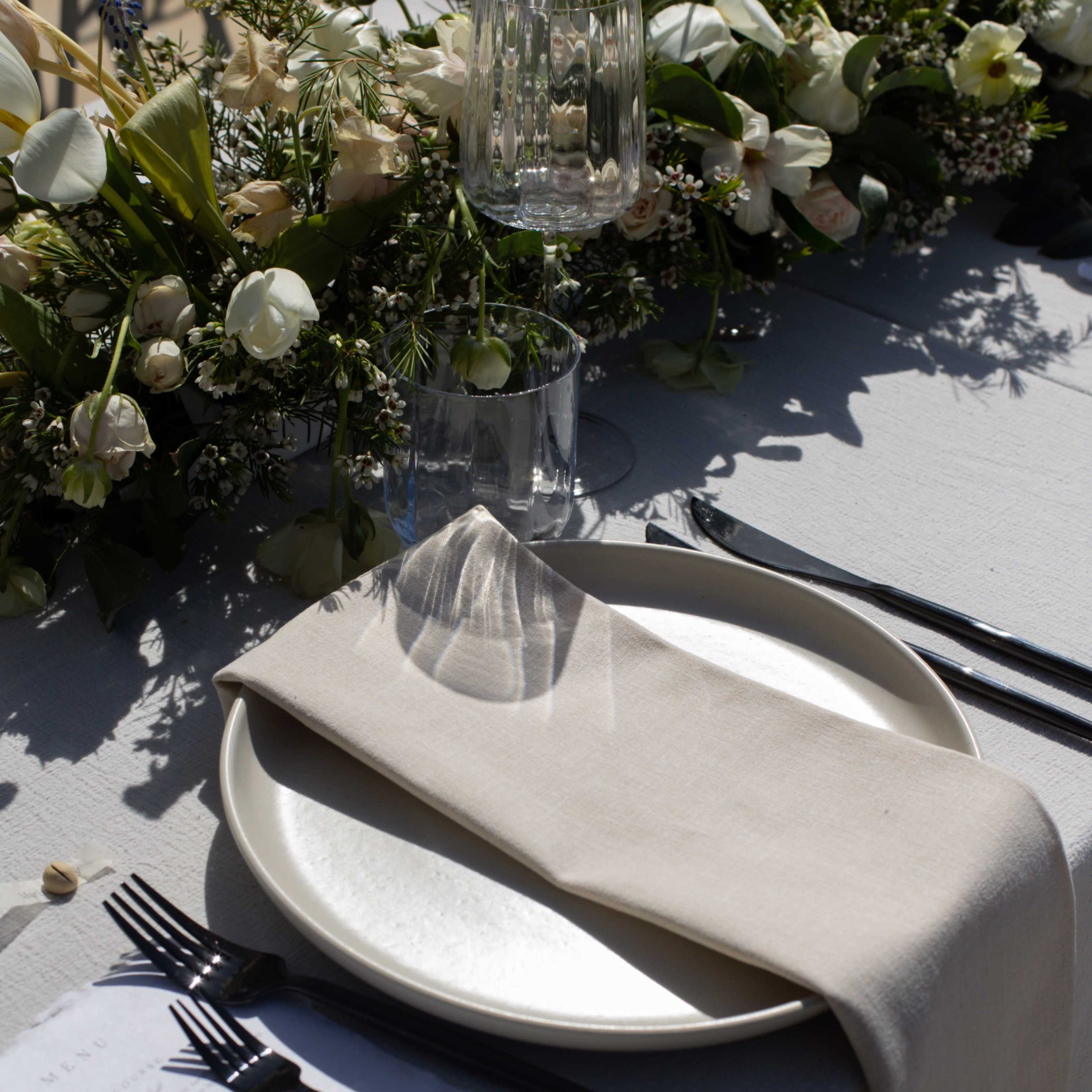 A dining table is set with a white plate, cloth napkin, silverware, and a menu, adorned with a floral arrangement.