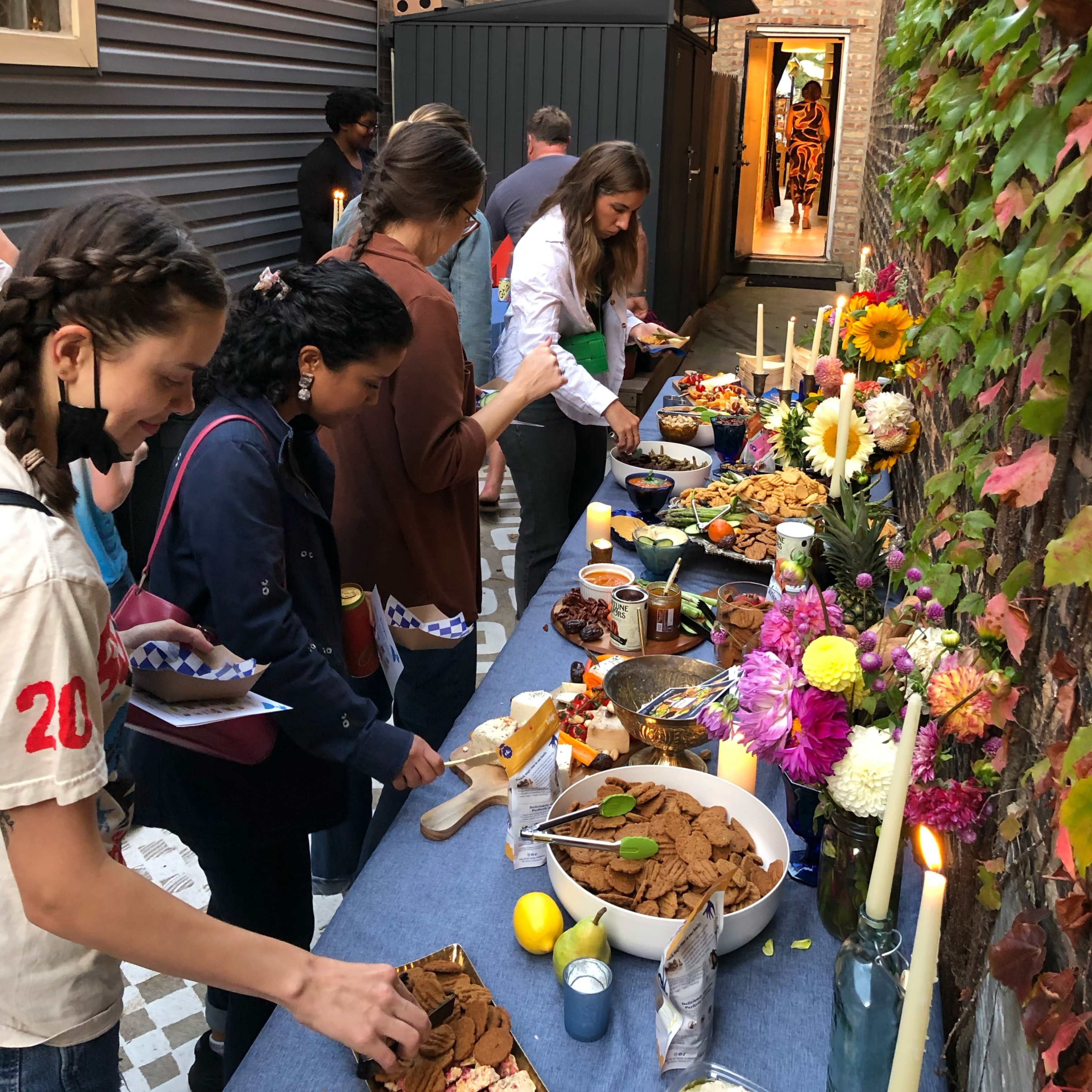 A group of people is serving food from a long table adorned with snacks and flowers in an outdoor alleyway lit with string lights.