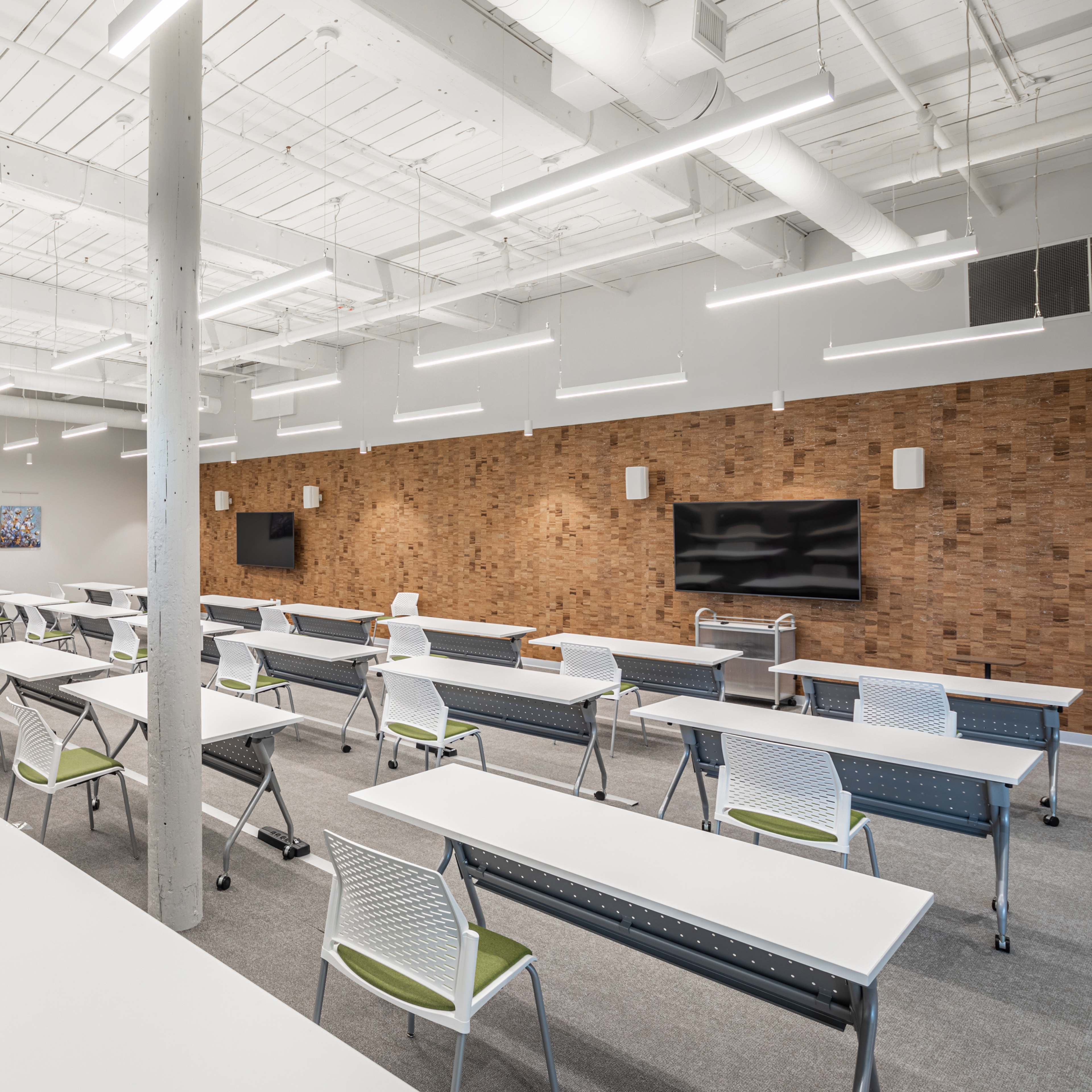 A brightly lit classroom features several rows of folding tables and chairs, with a large television displayed on a wooden accent wall.