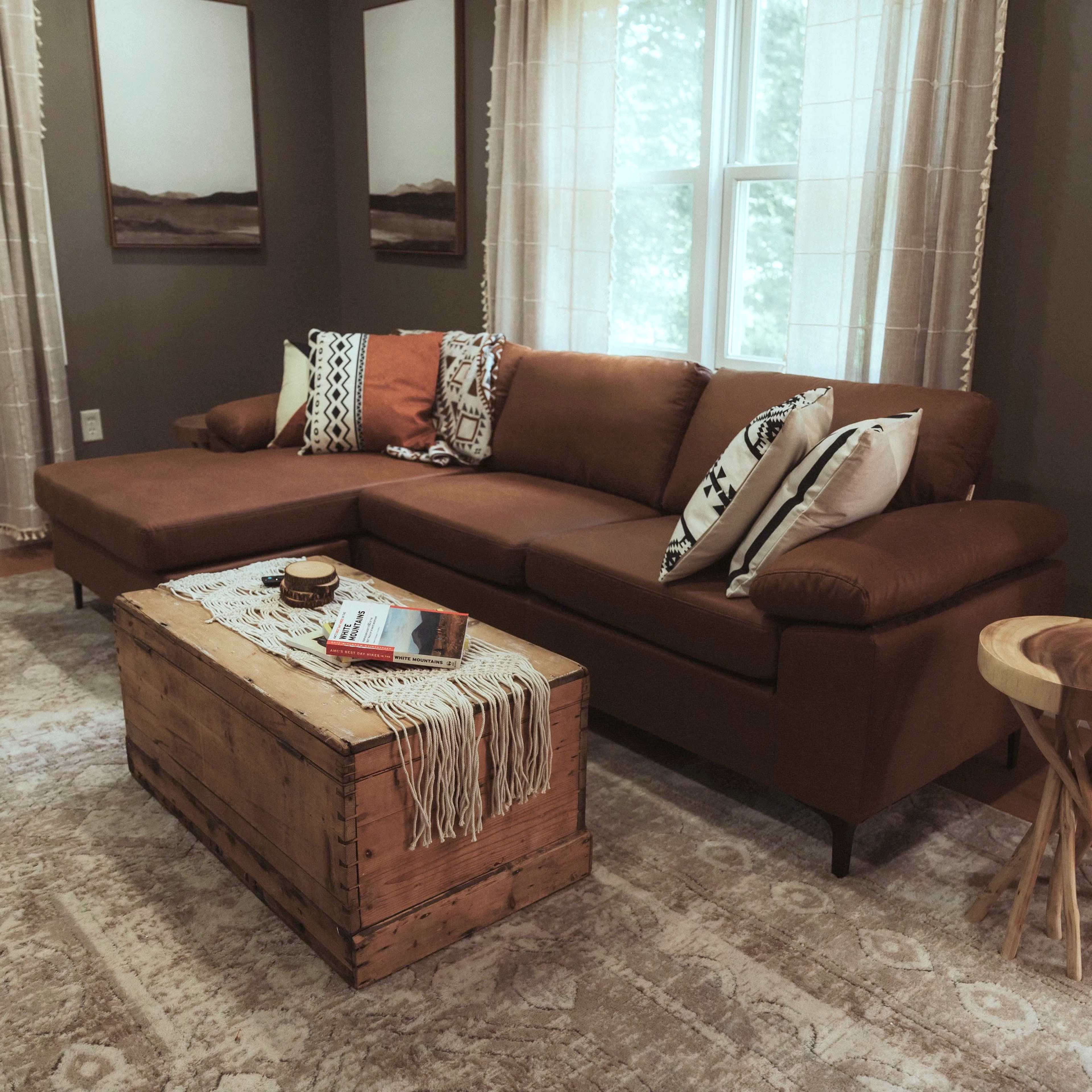 A brown sectional sofa with decorative pillows, a wooden coffee table, and a patterned rug, set in a living room with large windows that allow natural light.