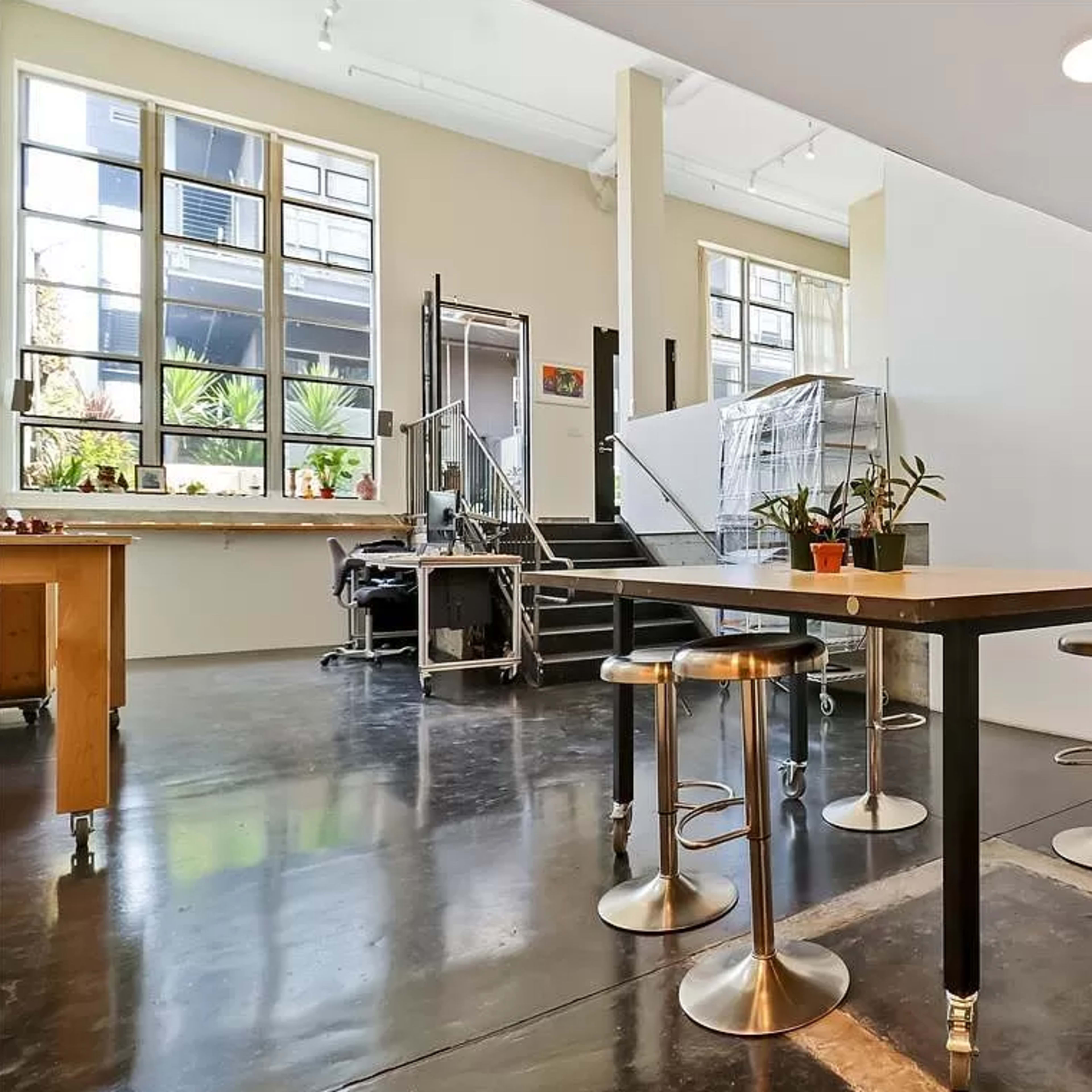 A modern interior space featuring an open layout with a table and stools, a large window letting in natural light, and a staircase in the background.