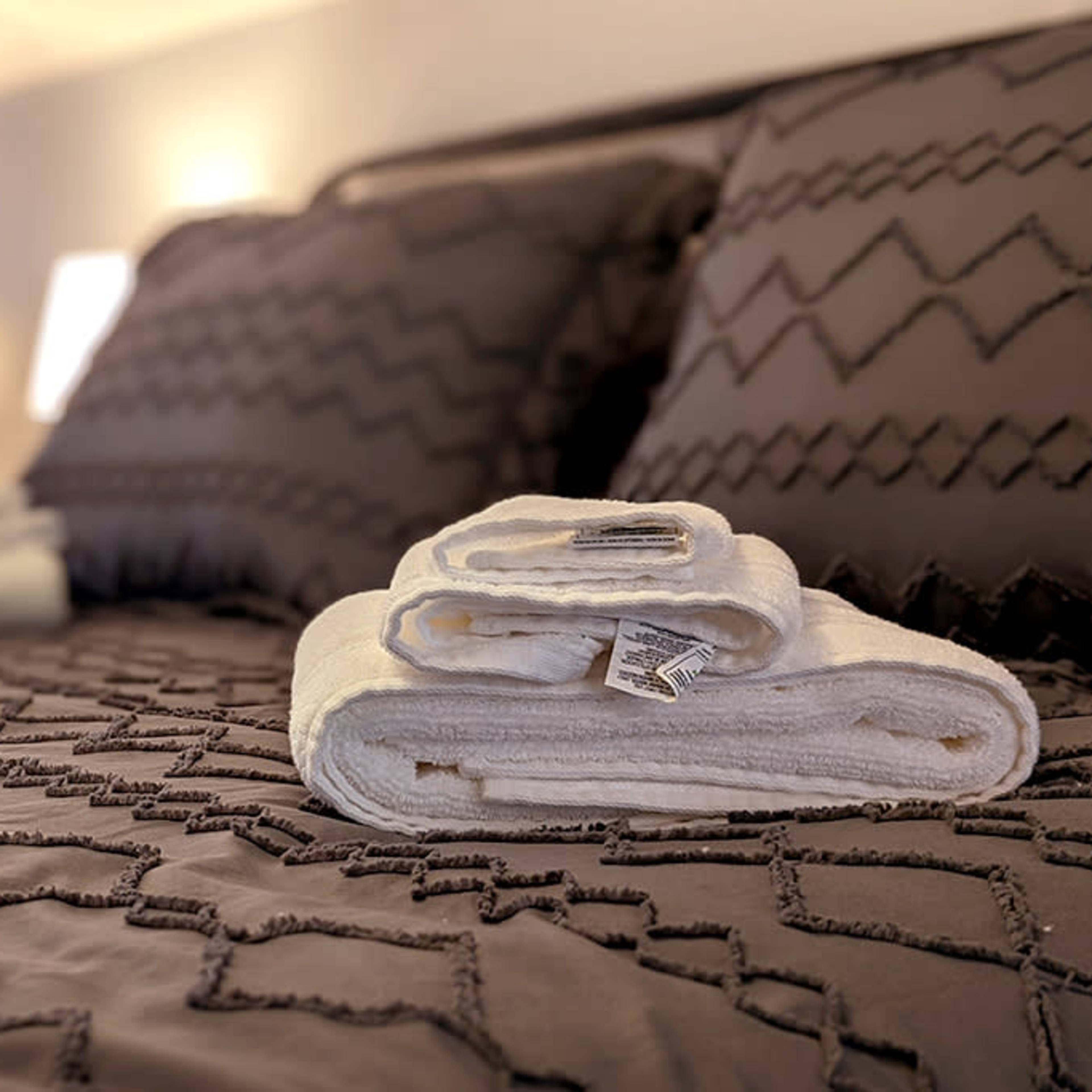A neatly arranged set of white towels is placed on a textured gray bedspread with two pillows in the background.