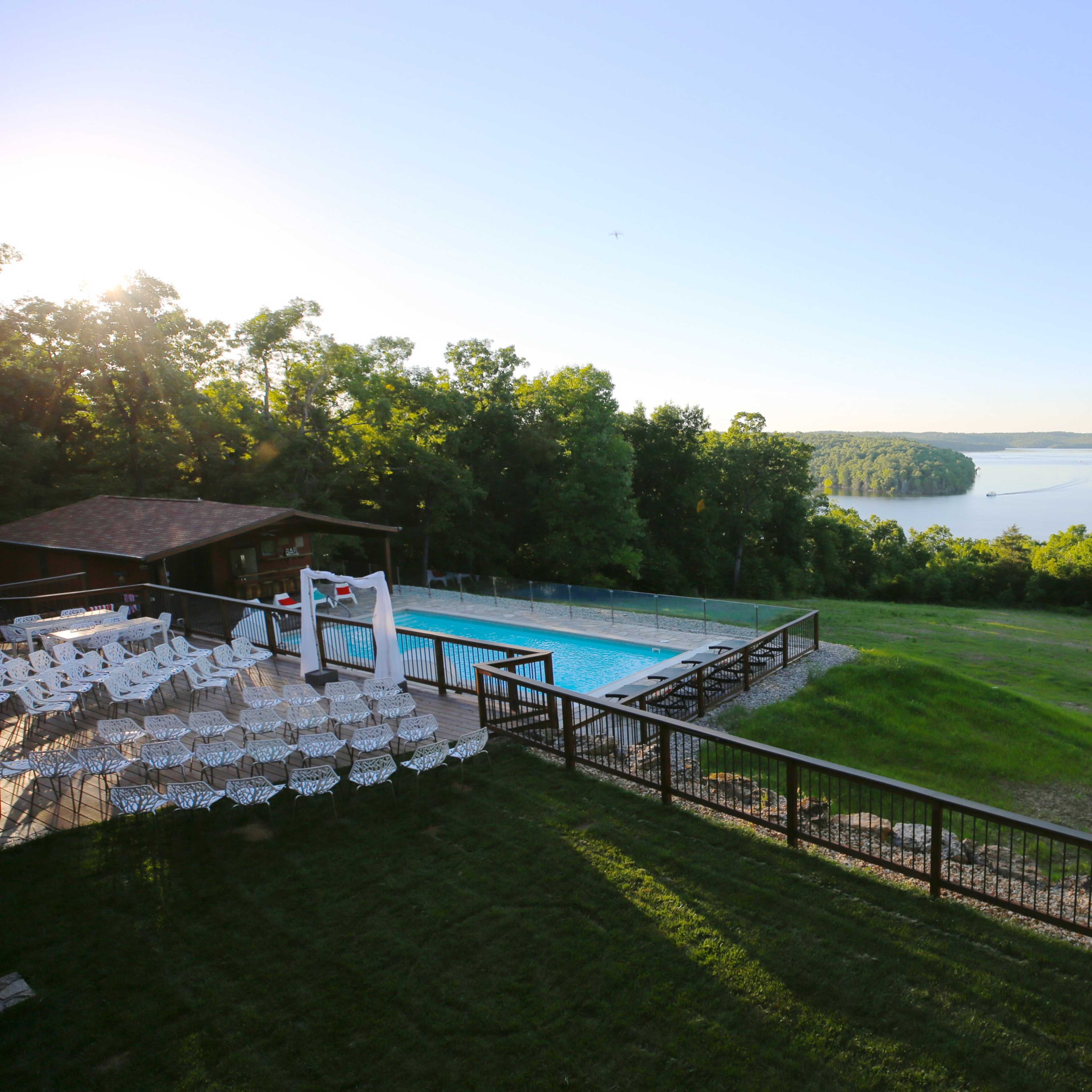 A swimming pool with lounge chairs overlooks a scenic lake surrounded by trees.