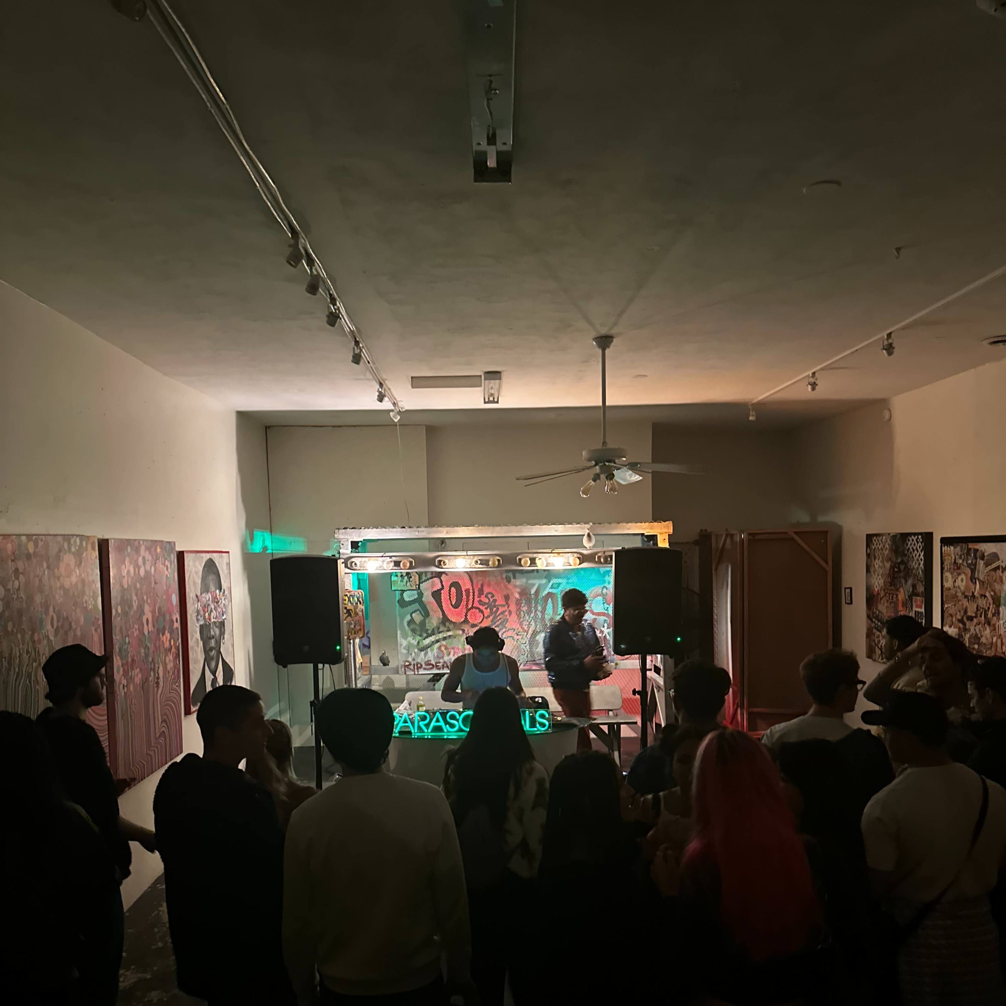 A crowd gathers in an art gallery as performers play music on a small stage with colorful lights.