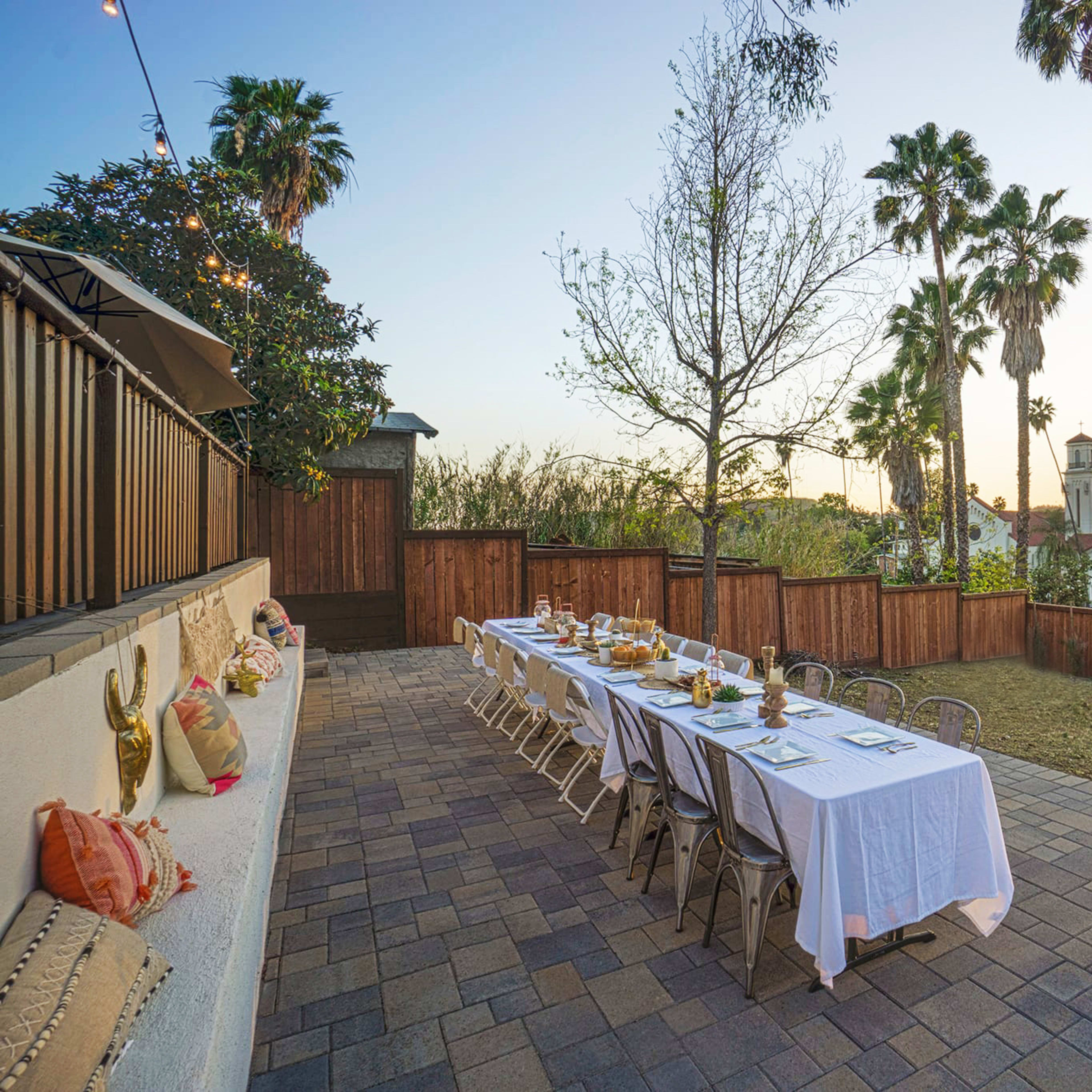 A long outdoor dining table is set with white linens and dinnerware, surrounded by palm trees at sunset.