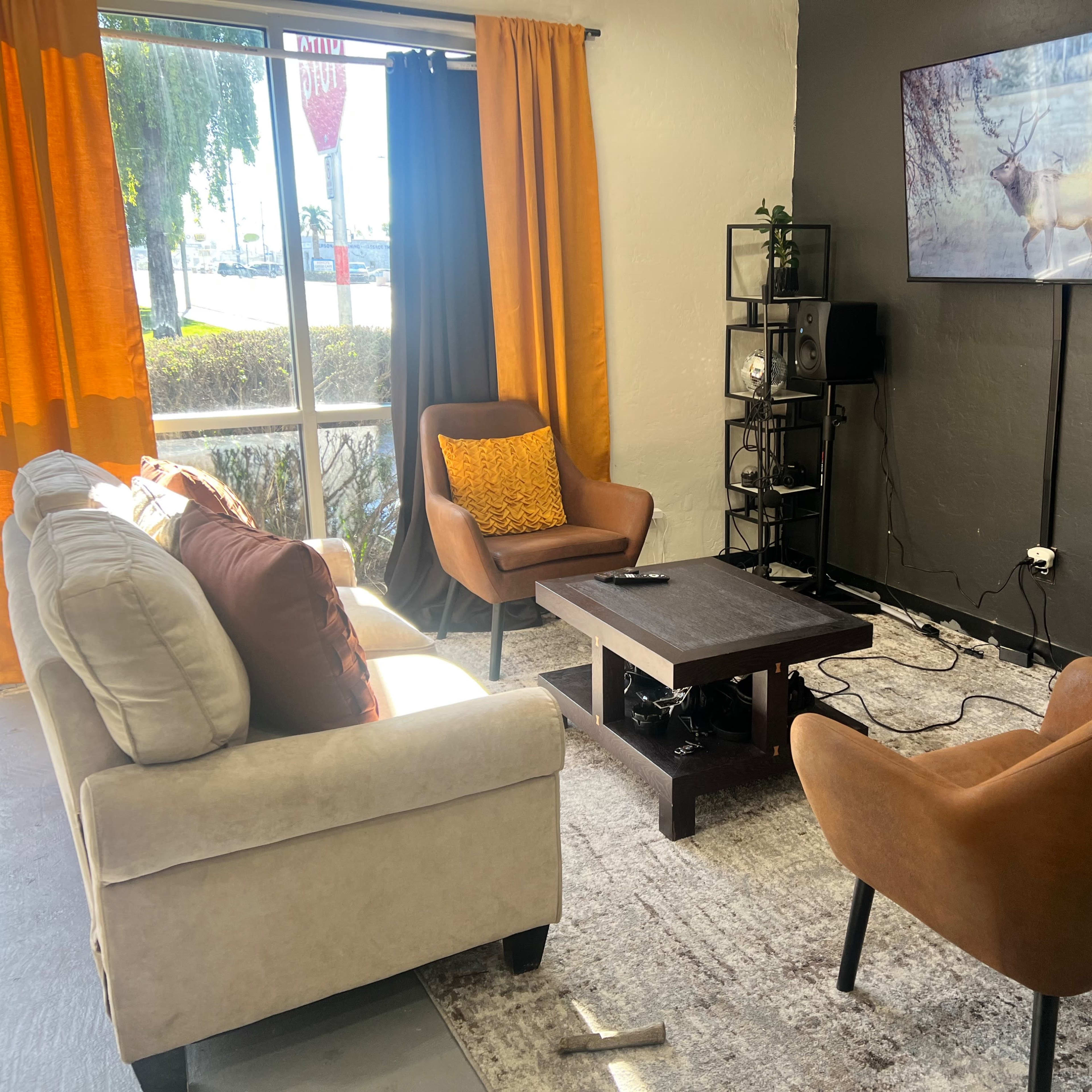 A cozy living room features two armchairs, a sofa, a coffee table, and a television mounted on the wall, with large windows letting in natural light.