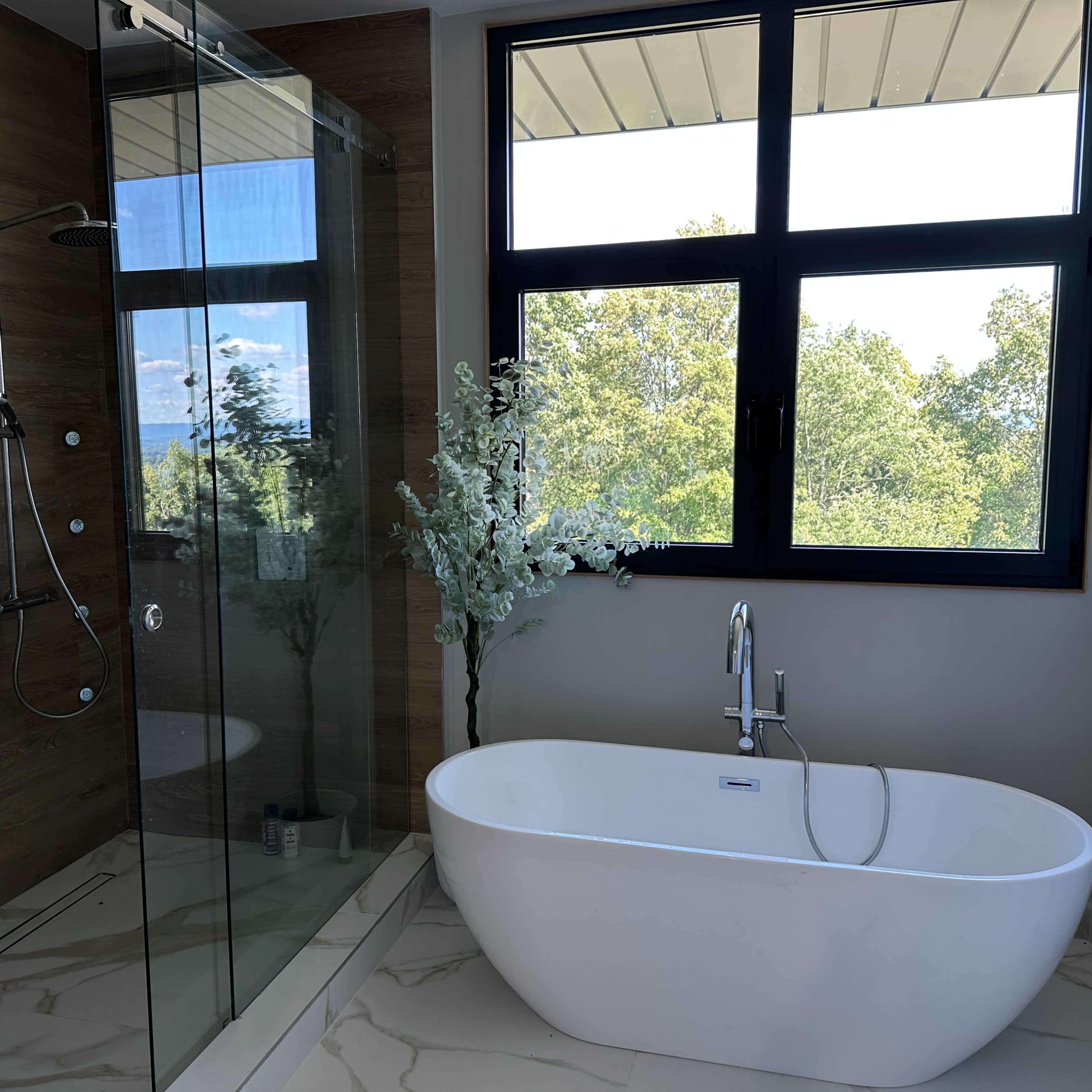A modern bathroom featuring a glass shower enclosure, a freestanding bathtub, and a large window with a view of greenery outside.