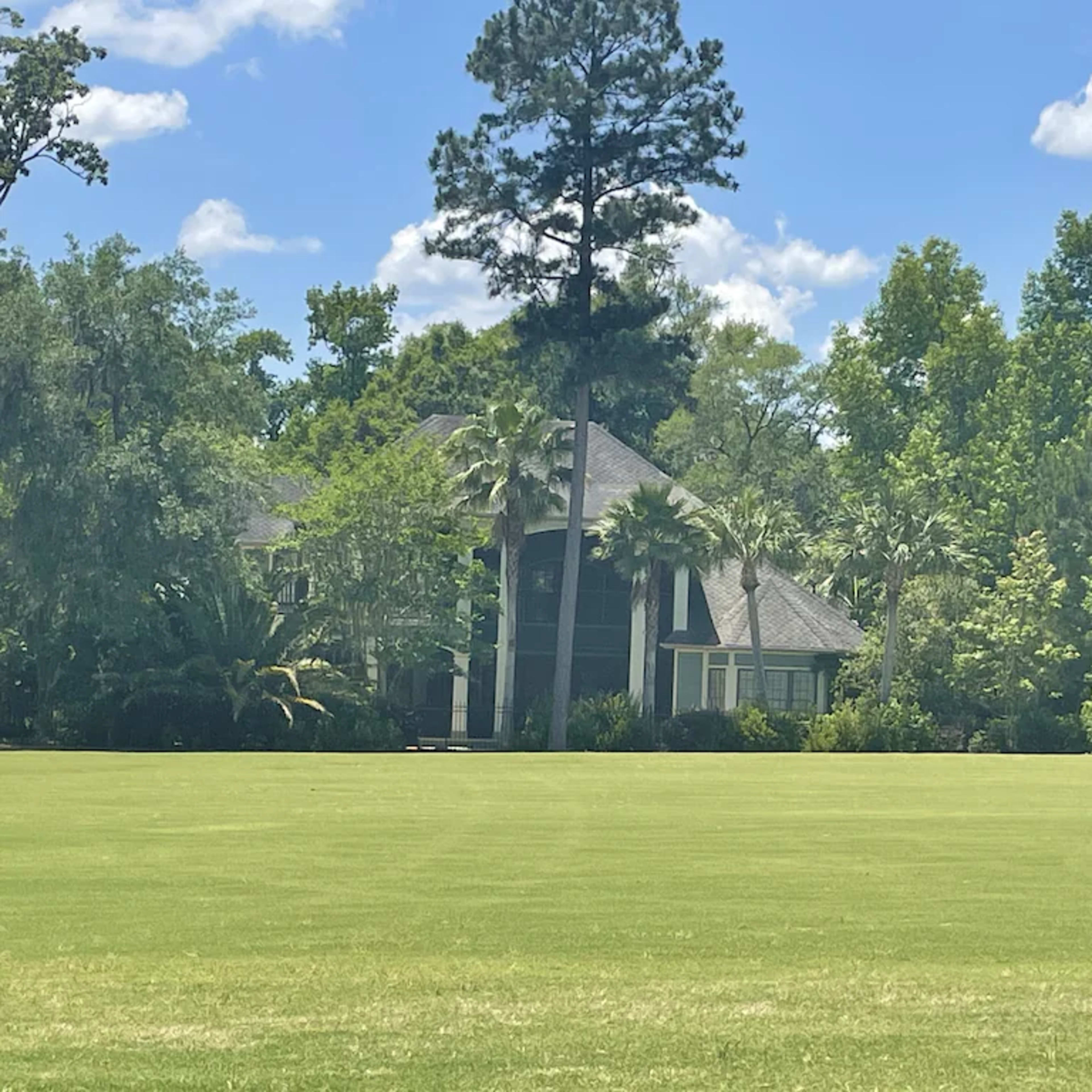 A large house stands surrounded by tall trees and lush greenery on a well-maintained lawn.