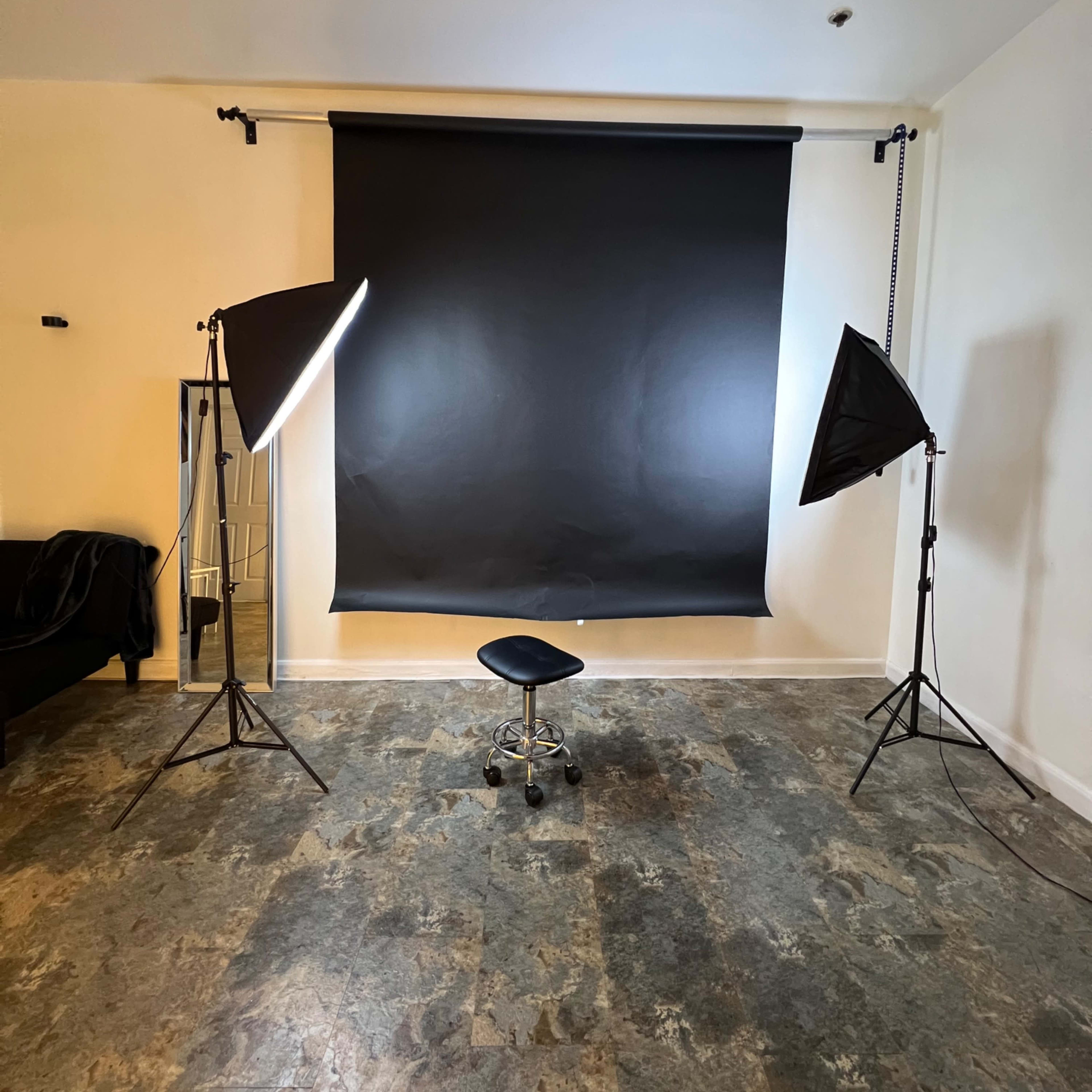 A photography studio setup with two umbrella lights, a black backdrop, and a black stool in the center.