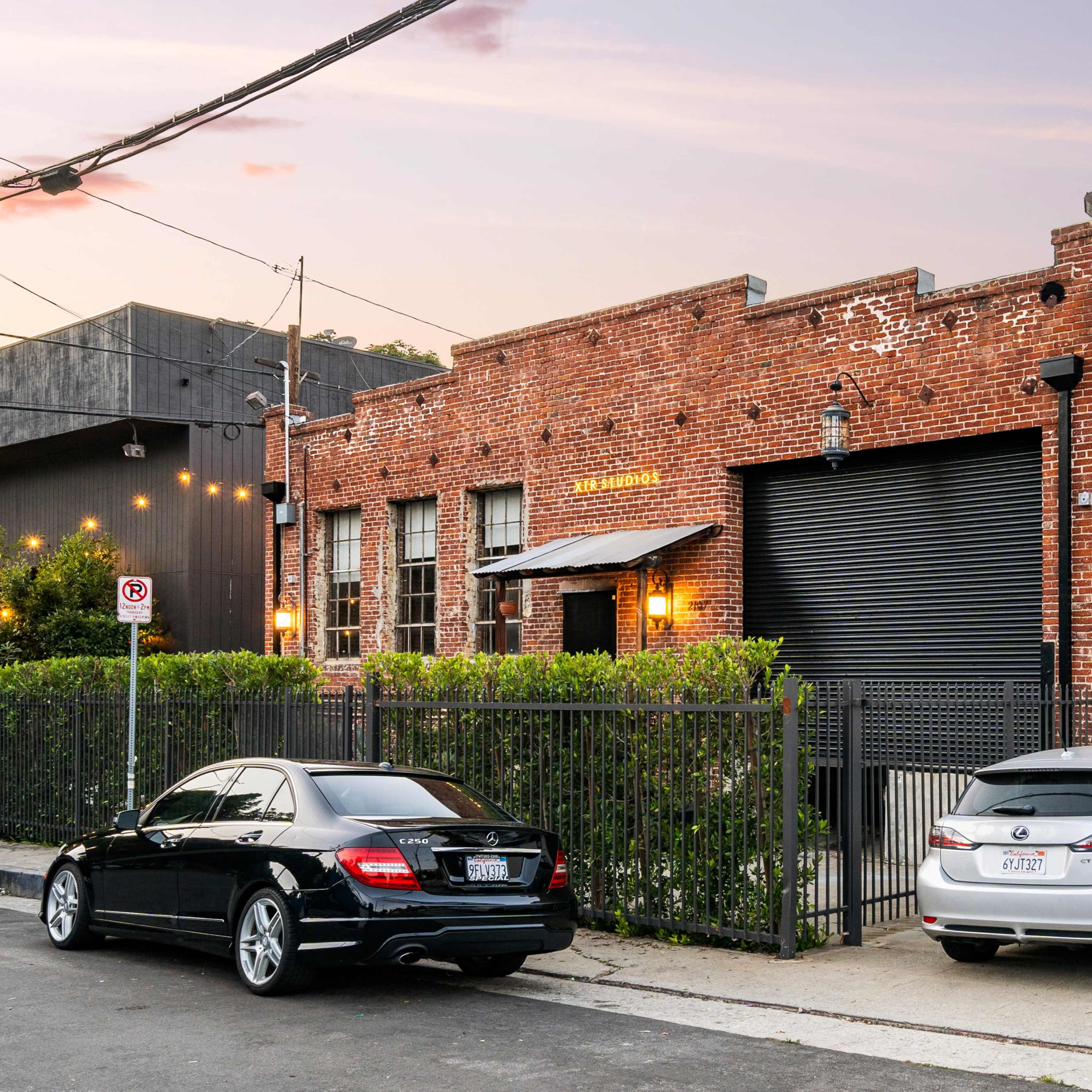 A brick building with a rolled-up black garage door, surrounded by parked cars along the street.