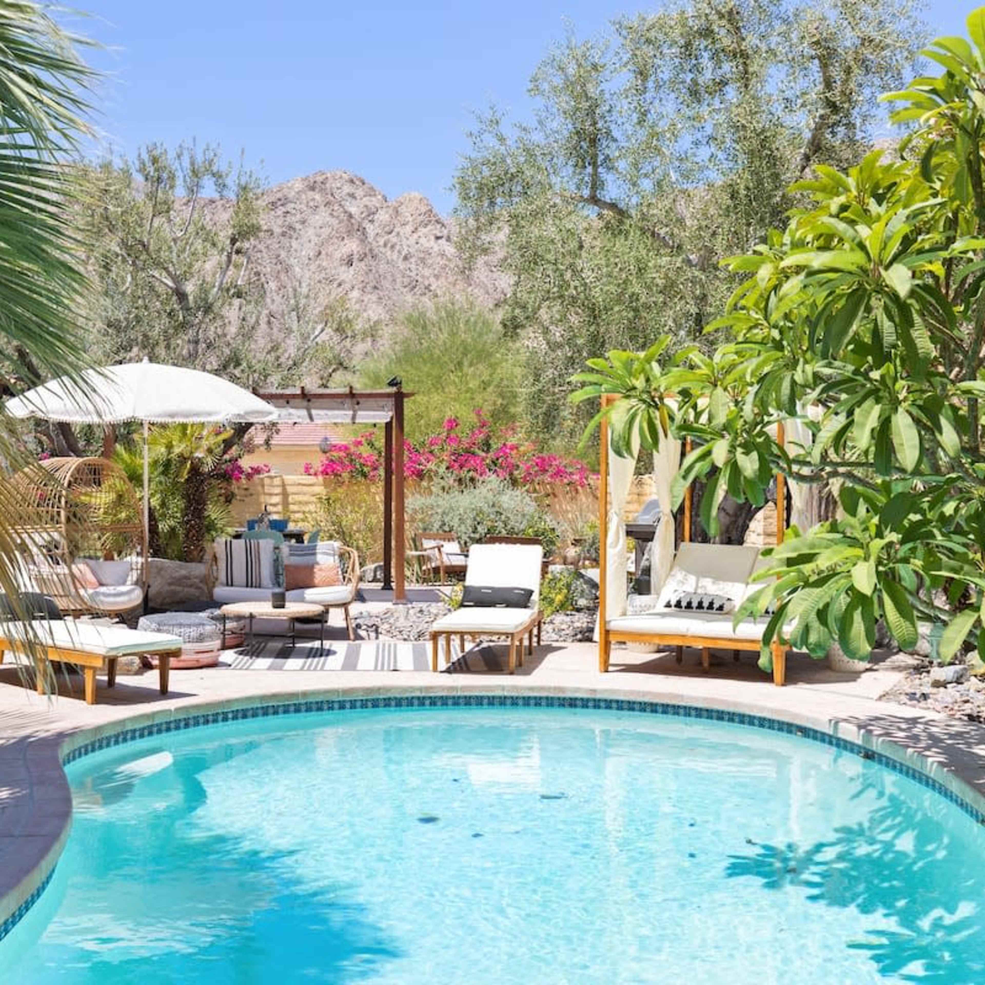 A backyard oasis features a turquoise pool surrounded by lounge chairs, umbrellas, and lush greenery set against a backdrop of mountains.
