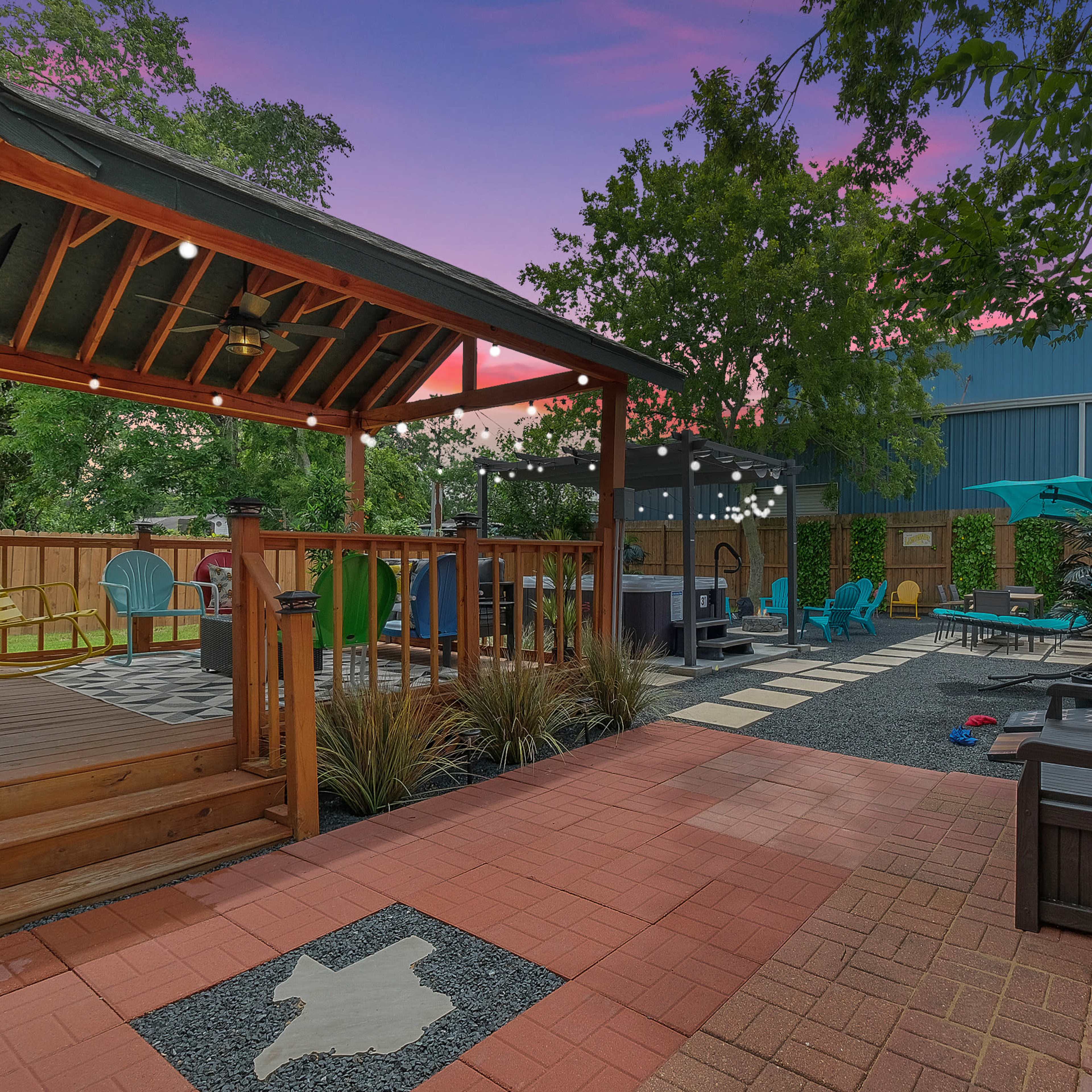 A patio area with wooden decking, colorful outdoor furniture, and a gazebo under evening light, surrounded by a wooden fence and green trees.