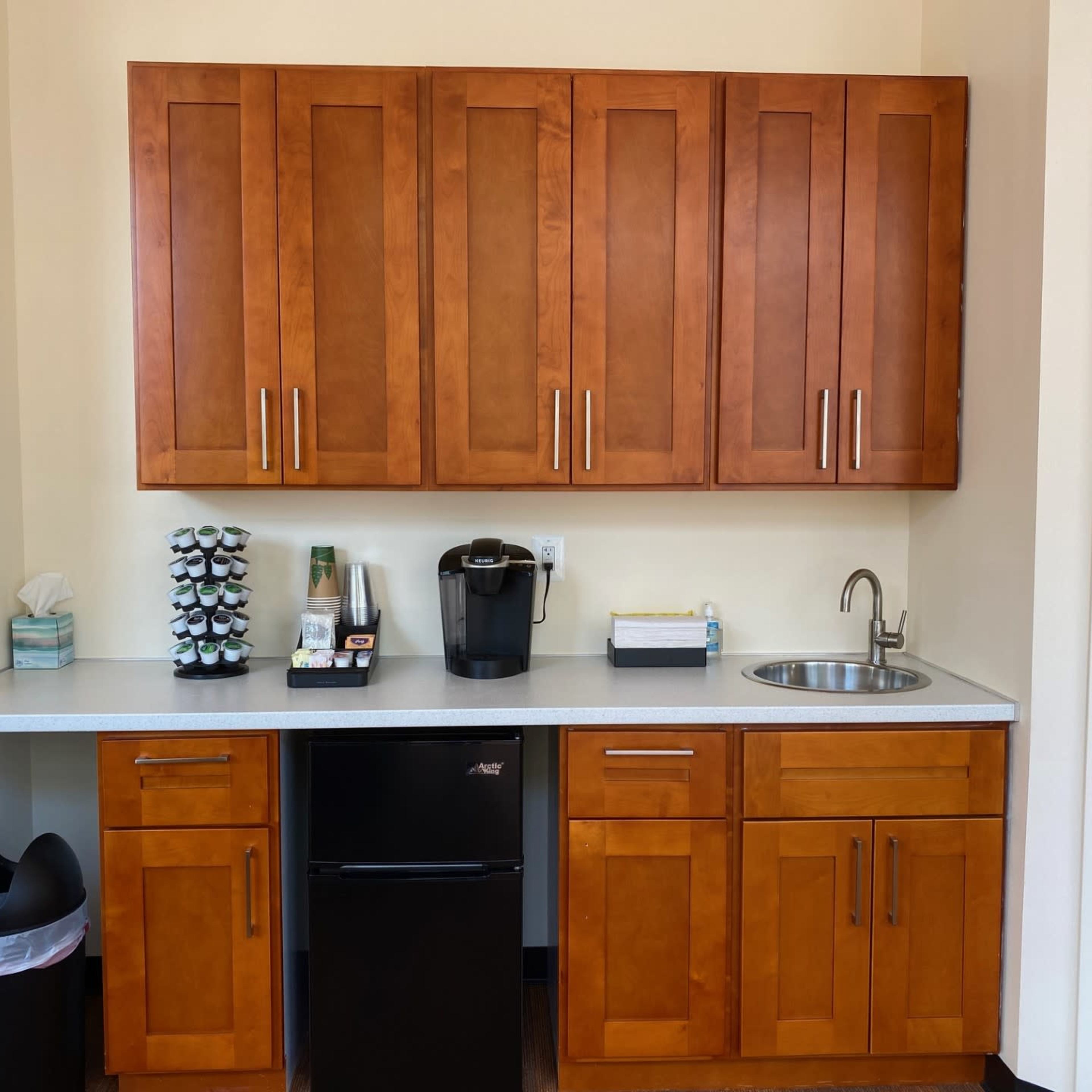A kitchenette with wooden cabinets, a countertop, a sink, a coffee maker, and a small refrigerator.
