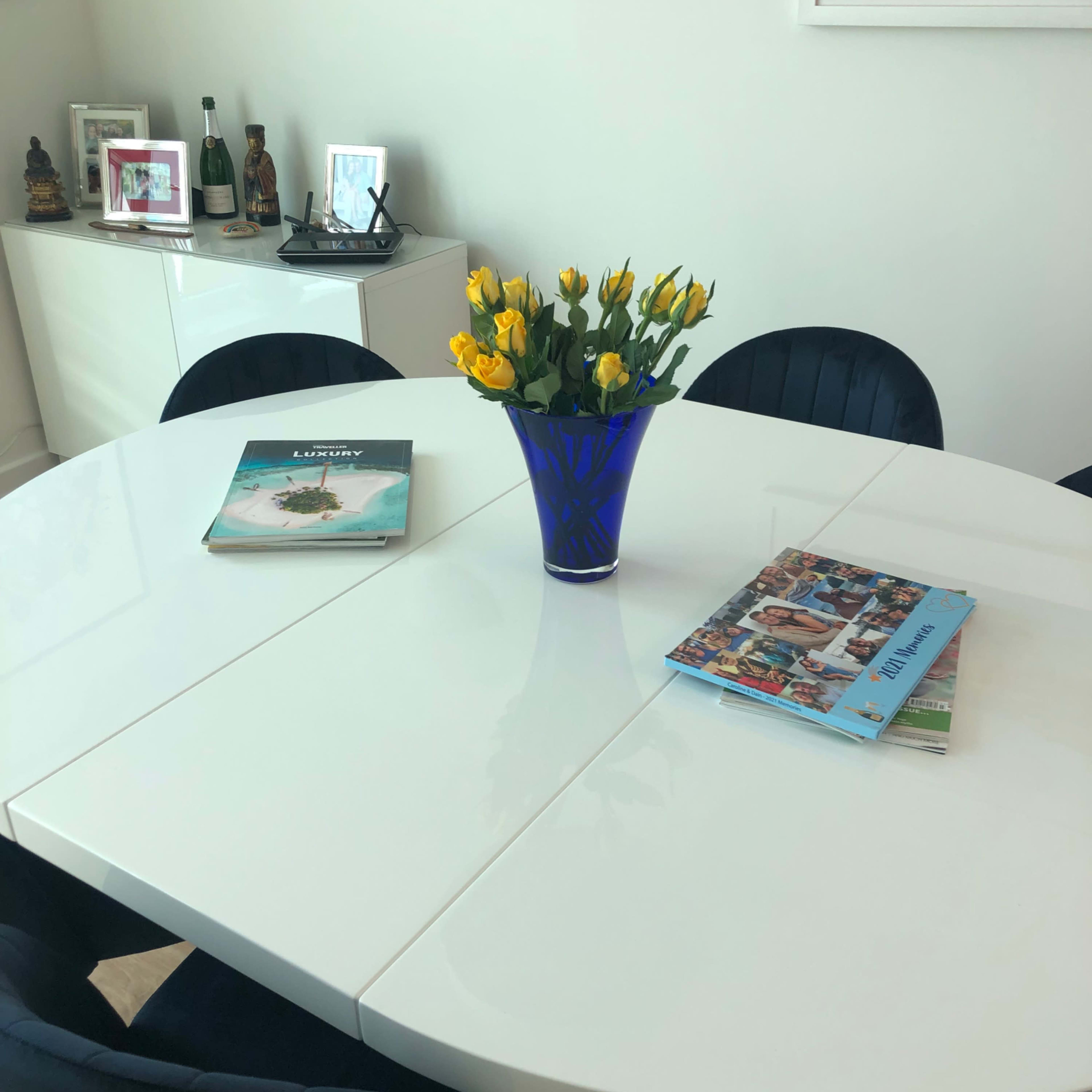 A white dining table is set with two magazines and a blue vase of yellow flowers in the center, surrounded by black chairs.
