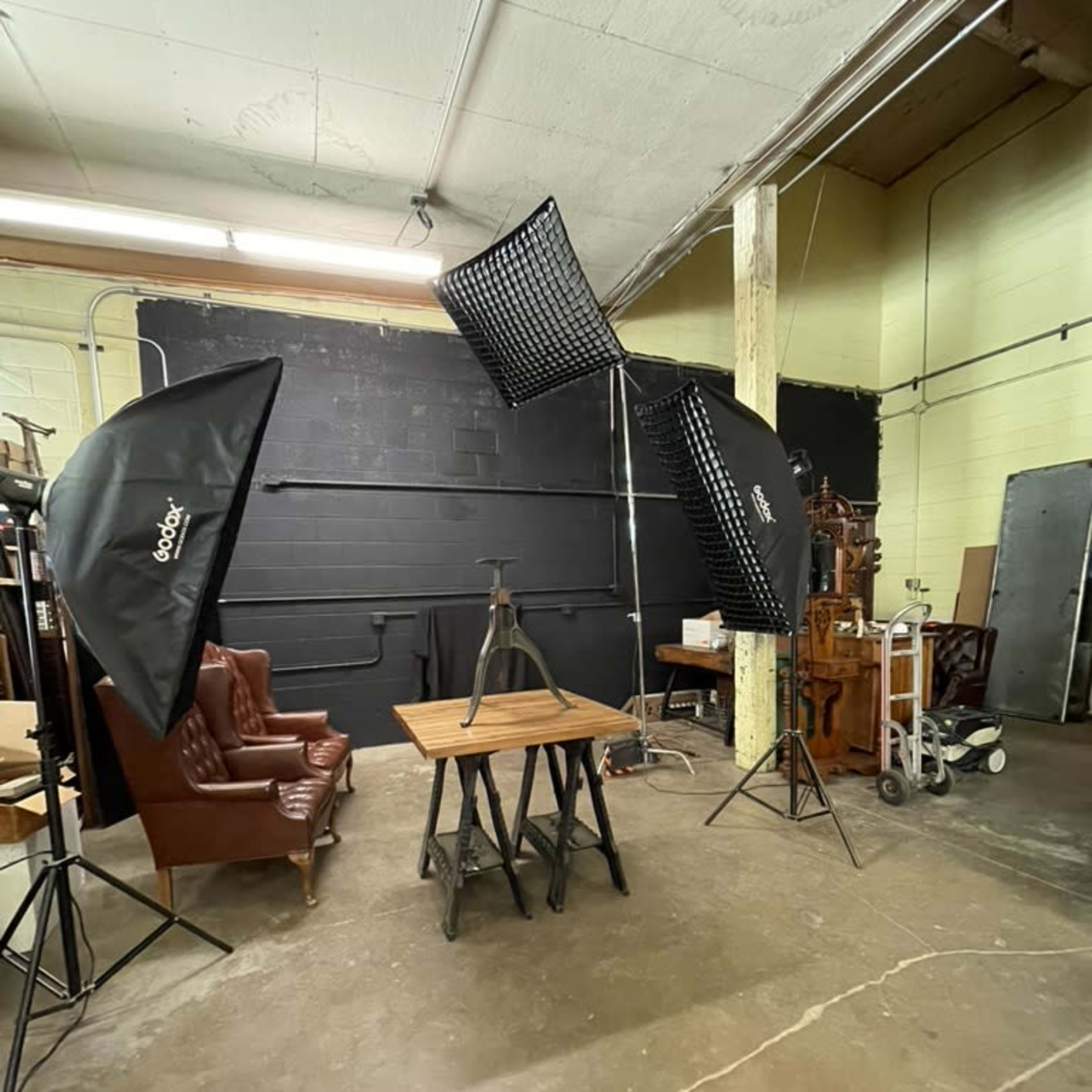 A photography studio setup with two large softbox lights, a wooden table on a metal stand, and a leather armchair in the background.