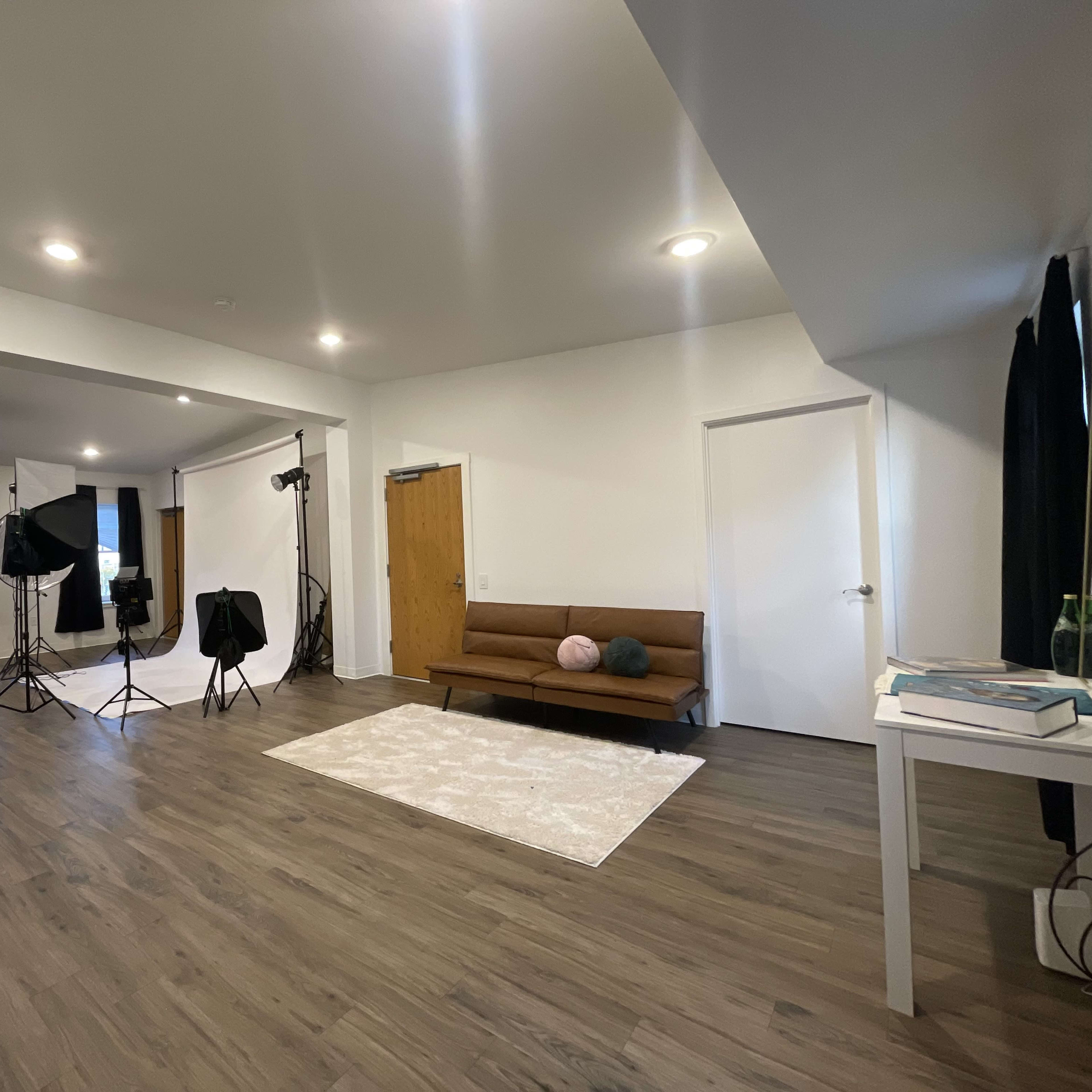 A spacious interior room featuring a brown sofa, lighting equipment, a rug, and large windows with black curtains.