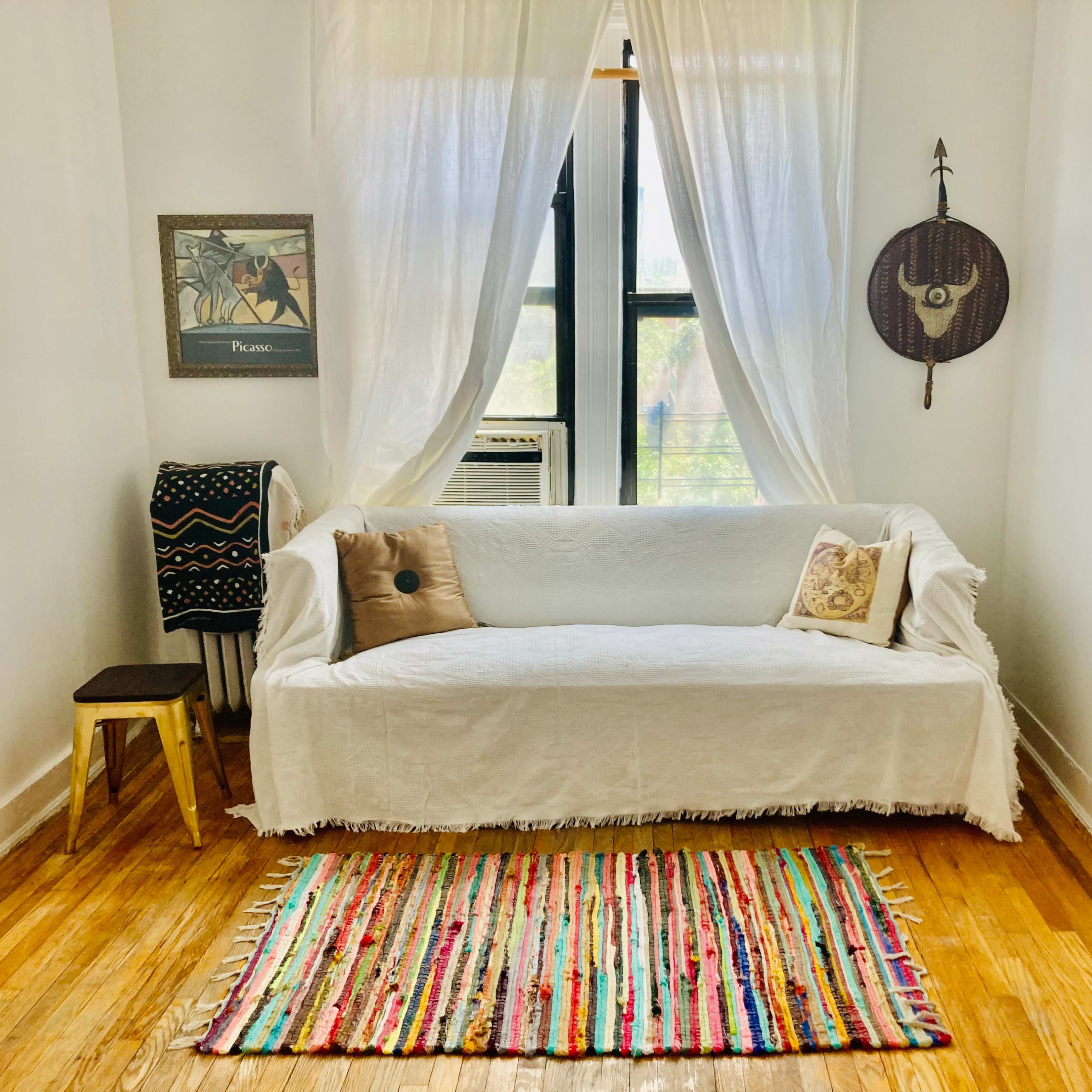 A white couch is draped with a textured covering and sits against a bright wall near a small wooden stool and a colorful woven rug.