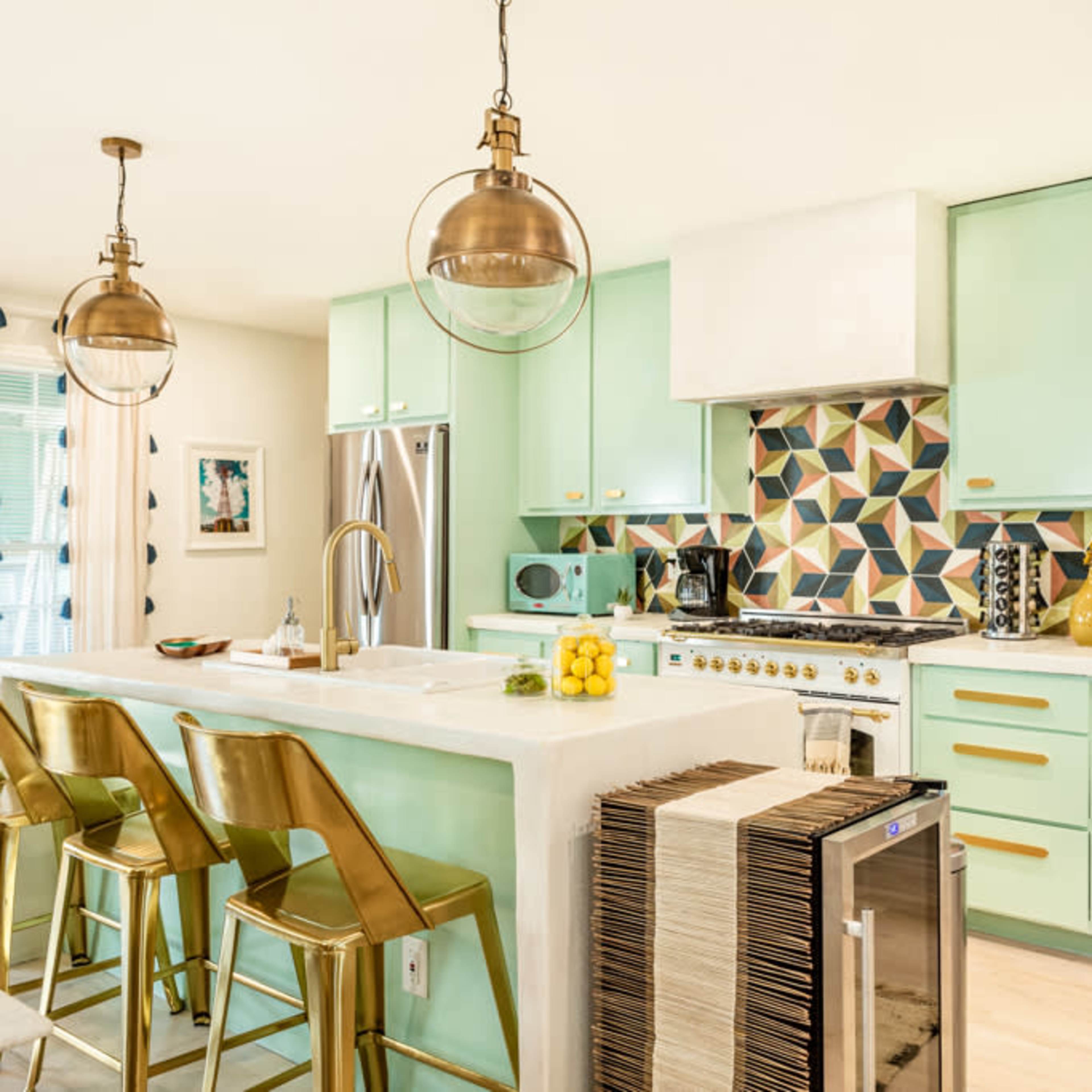 A modern kitchen features mint green cabinetry, a geometric tiled backsplash, and brass-accented bar stools around a white island.