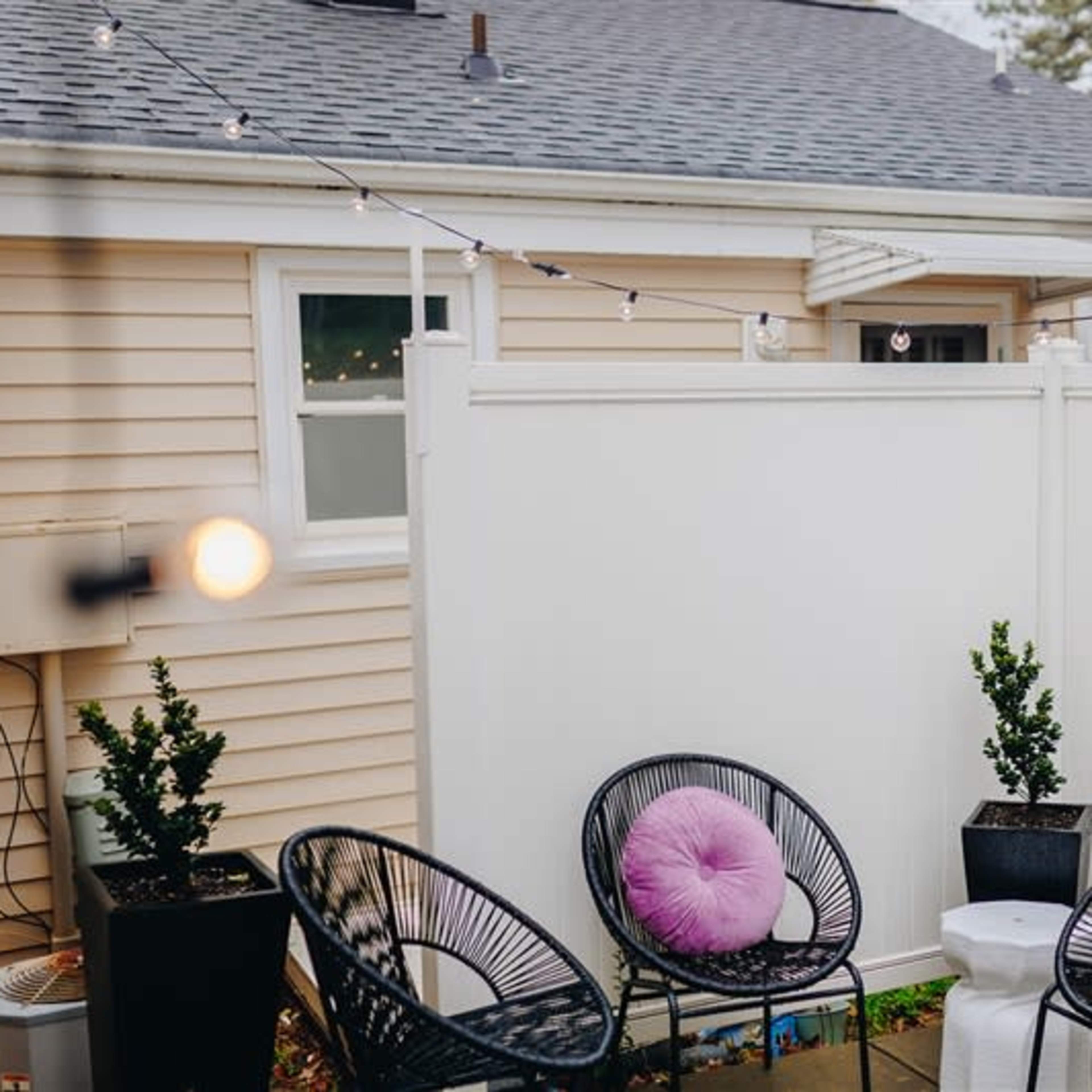 A small outdoor seating area features four black chairs with purple cushions arranged around a white side table, enclosed by a white fence with string lights above.