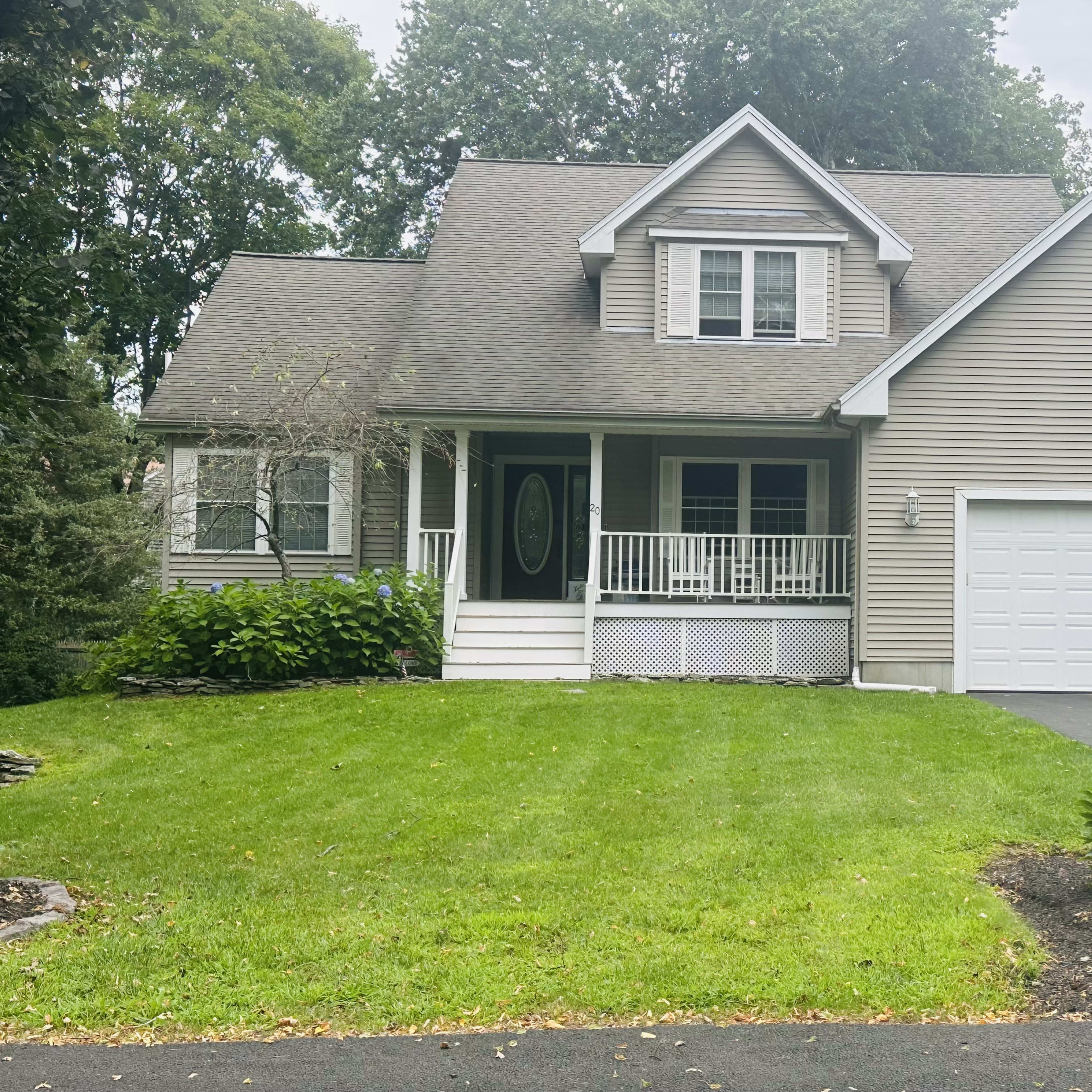 A two-story house with a peaked roof, front porch, and attached garage is situated on a green lawn surrounded by trees.