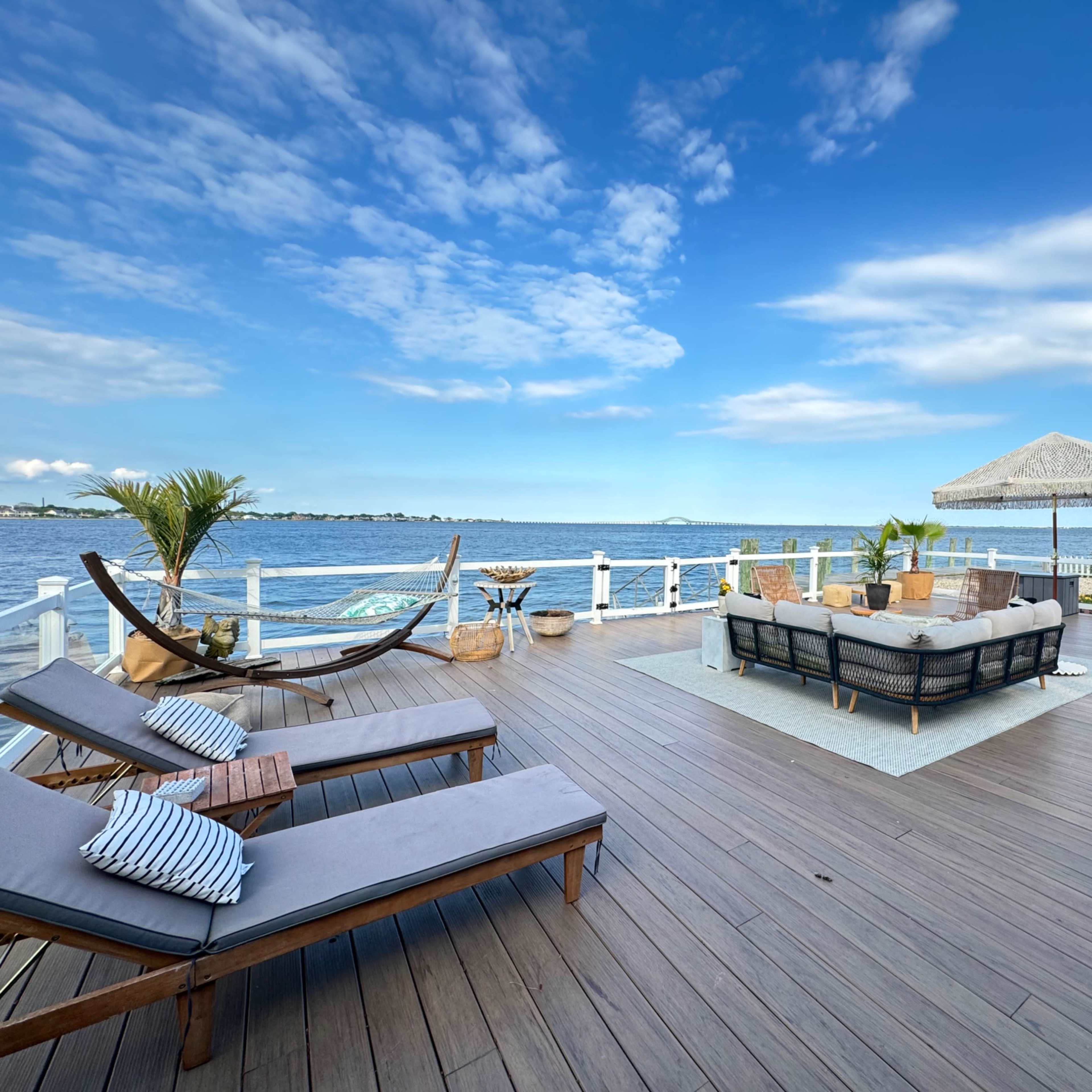 A tranquil outdoor deck overlooking a calm body of water, furnished with lounge chairs, a seating area, and a shaded gazebo.