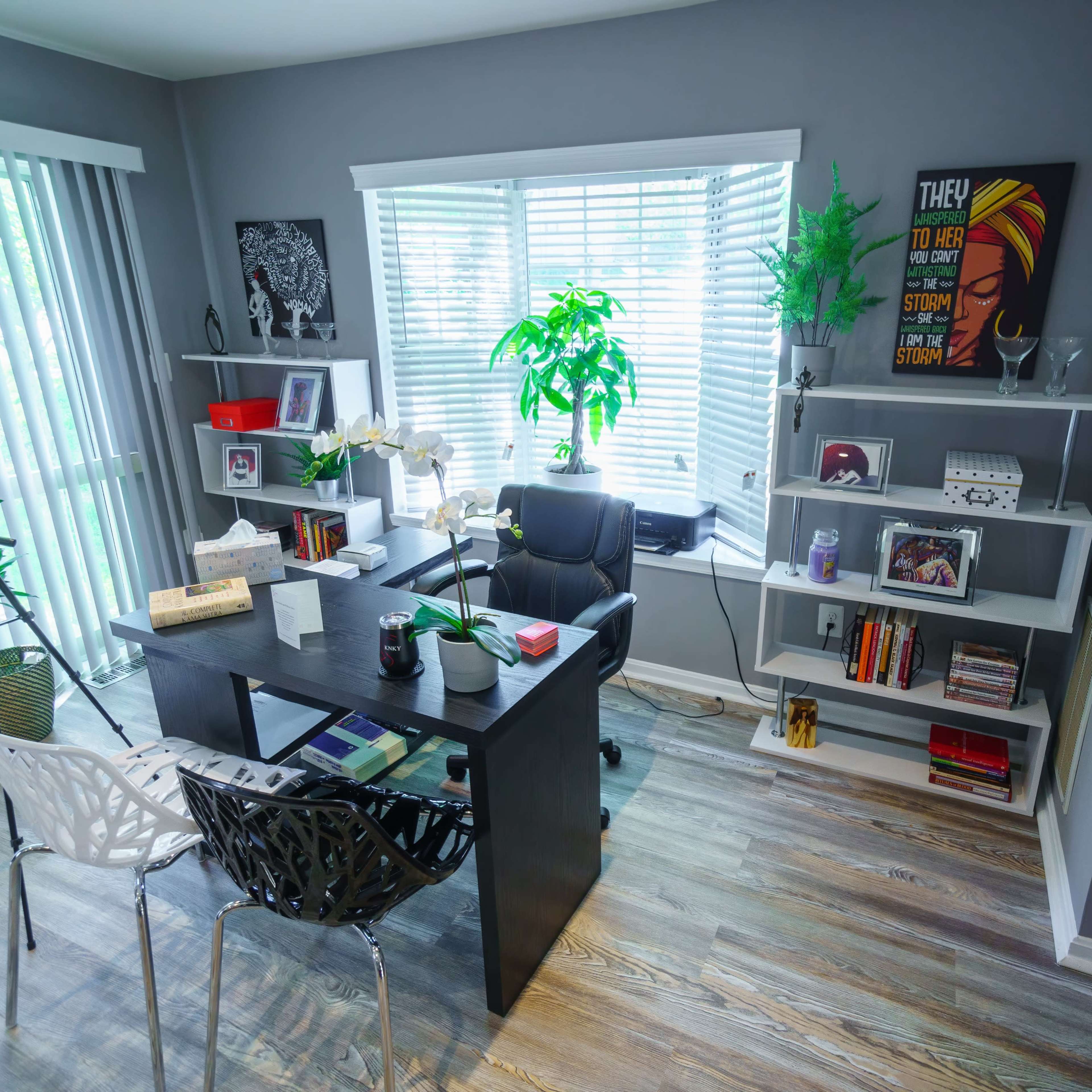 A modern office space features a black desk, shelves with decor and books, a chair, and a large window with blinds, allowing natural light to enter.