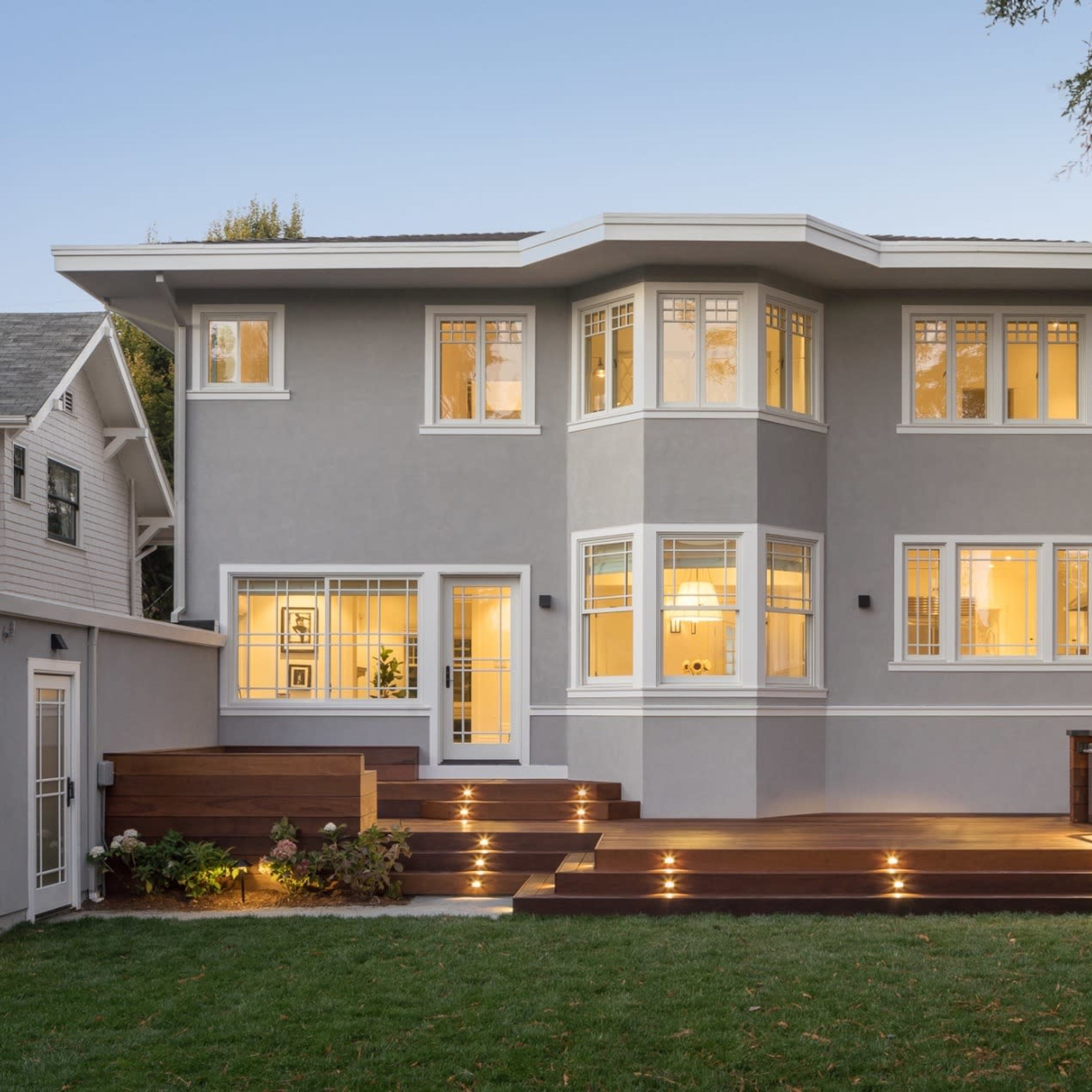 A modern two-story house with a gray exterior, large windows, and a wooden deck, surrounded by a green lawn and trees.