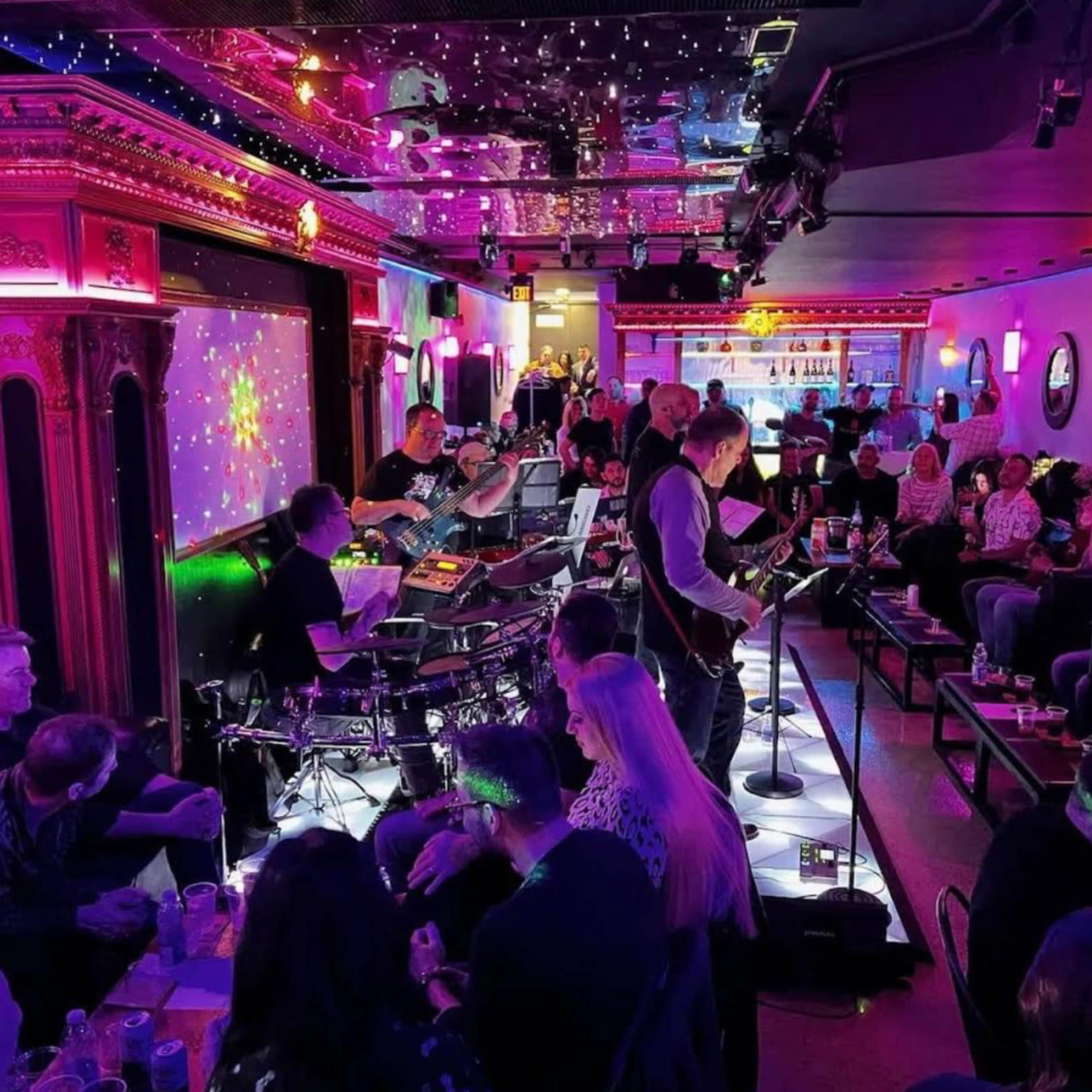 A live band performs on a stage in a brightly lit bar, while an audience sits at tables enjoying drinks.