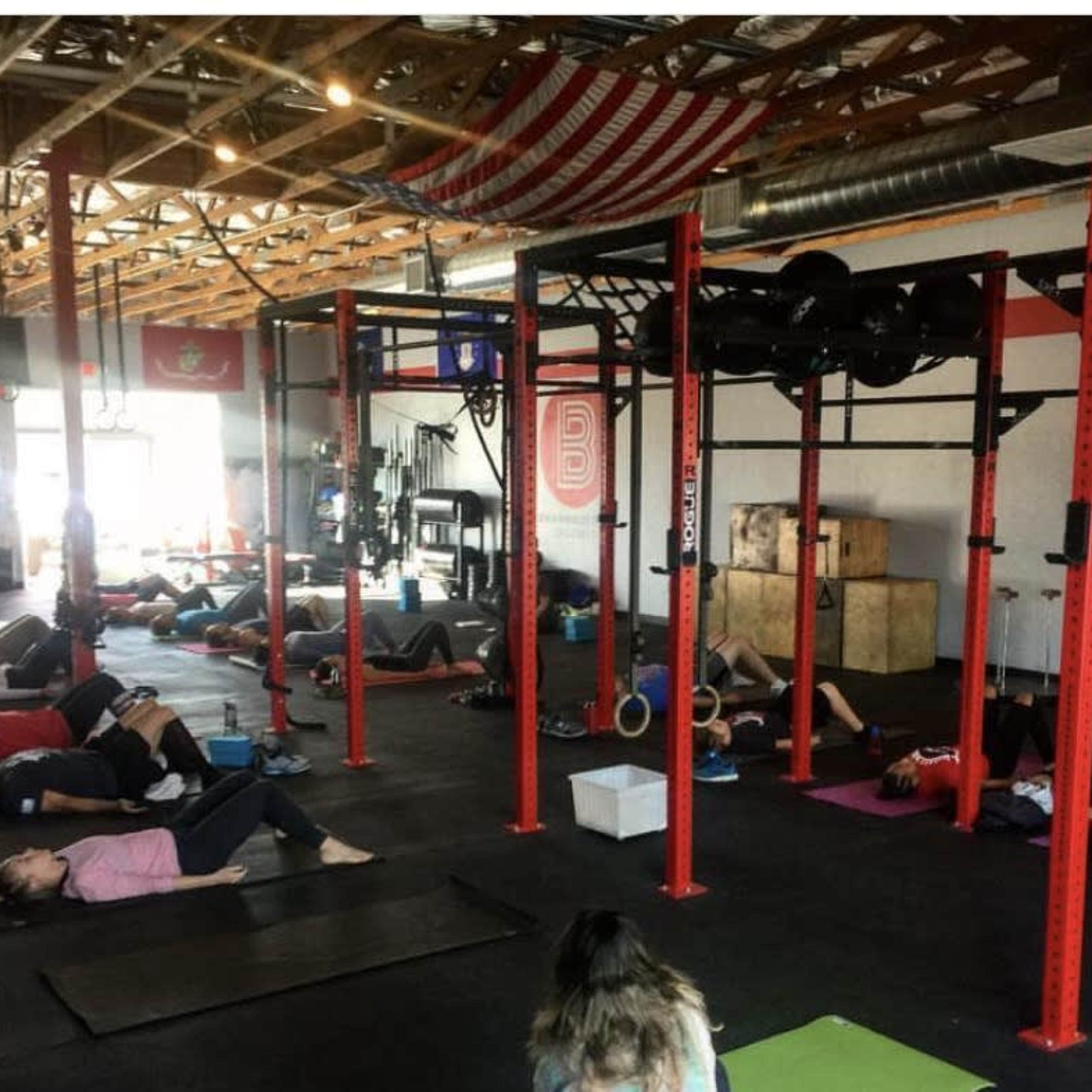 A group of people are lying on yoga mats in a gym, with fitness equipment and an American flag visible in the background.