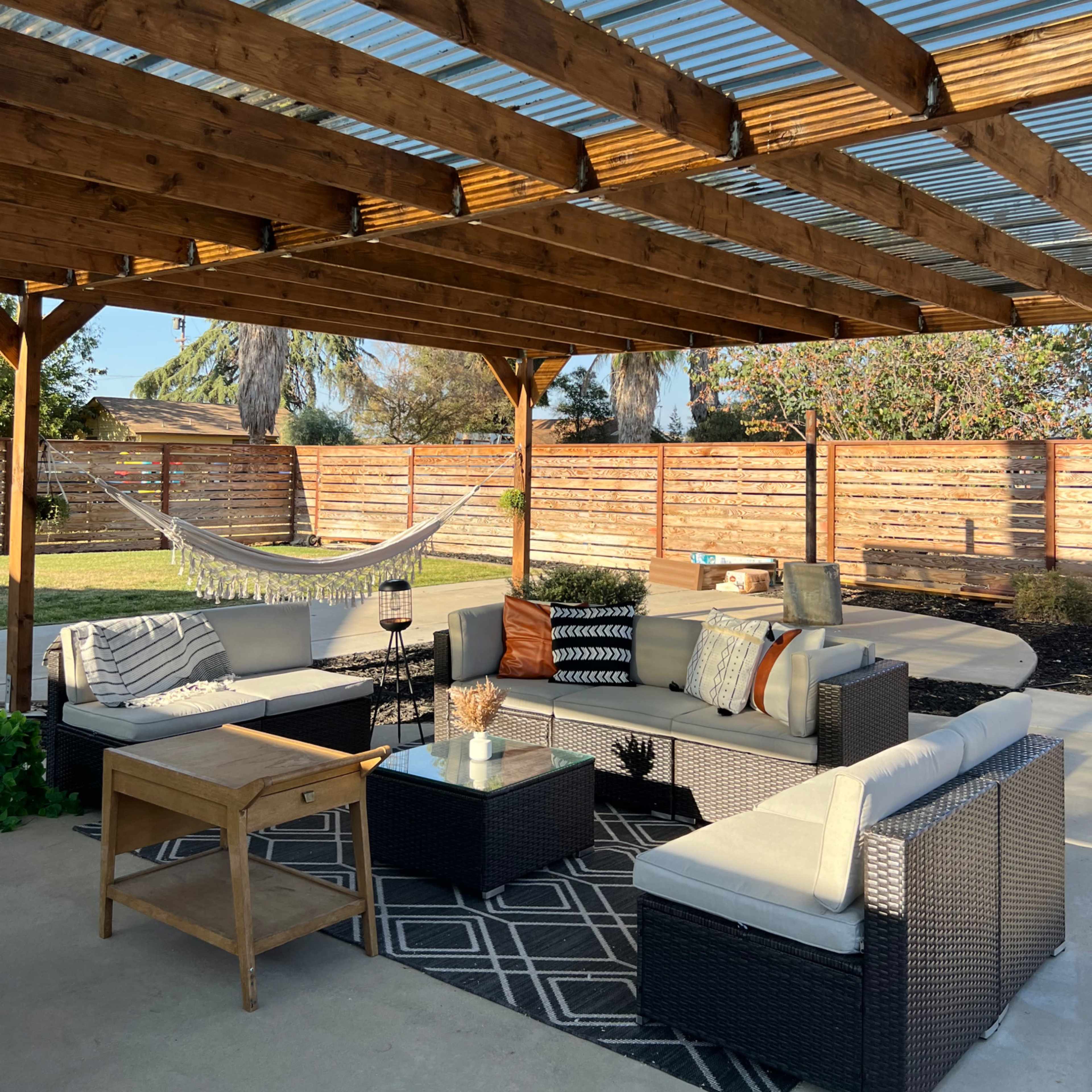 A covered outdoor seating area features a sectional sofa, a coffee table, and a hammock surrounded by a green yard and wooden fencing.