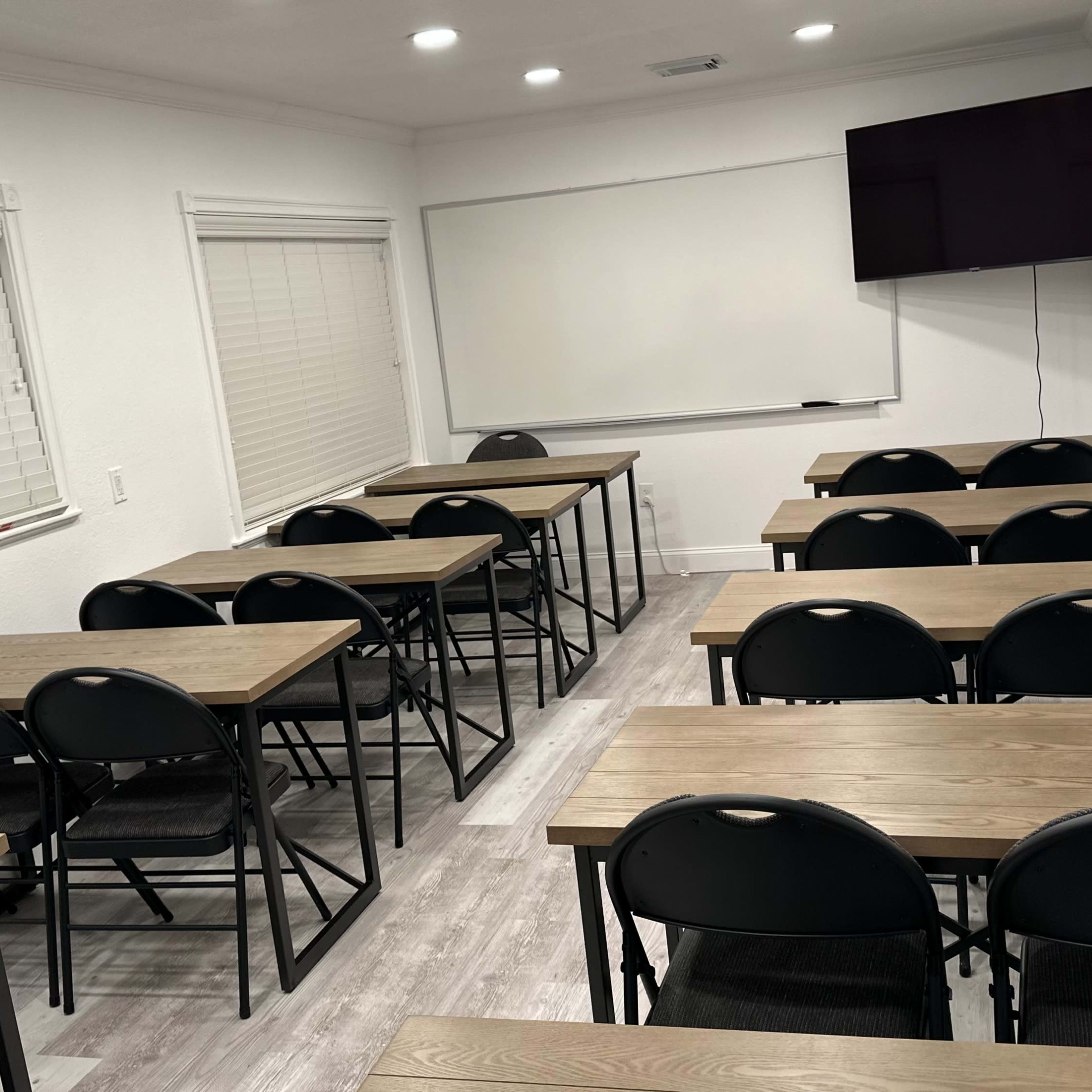 A classroom with several rows of tables and chairs arranged facing a whiteboard and a wall-mounted television.