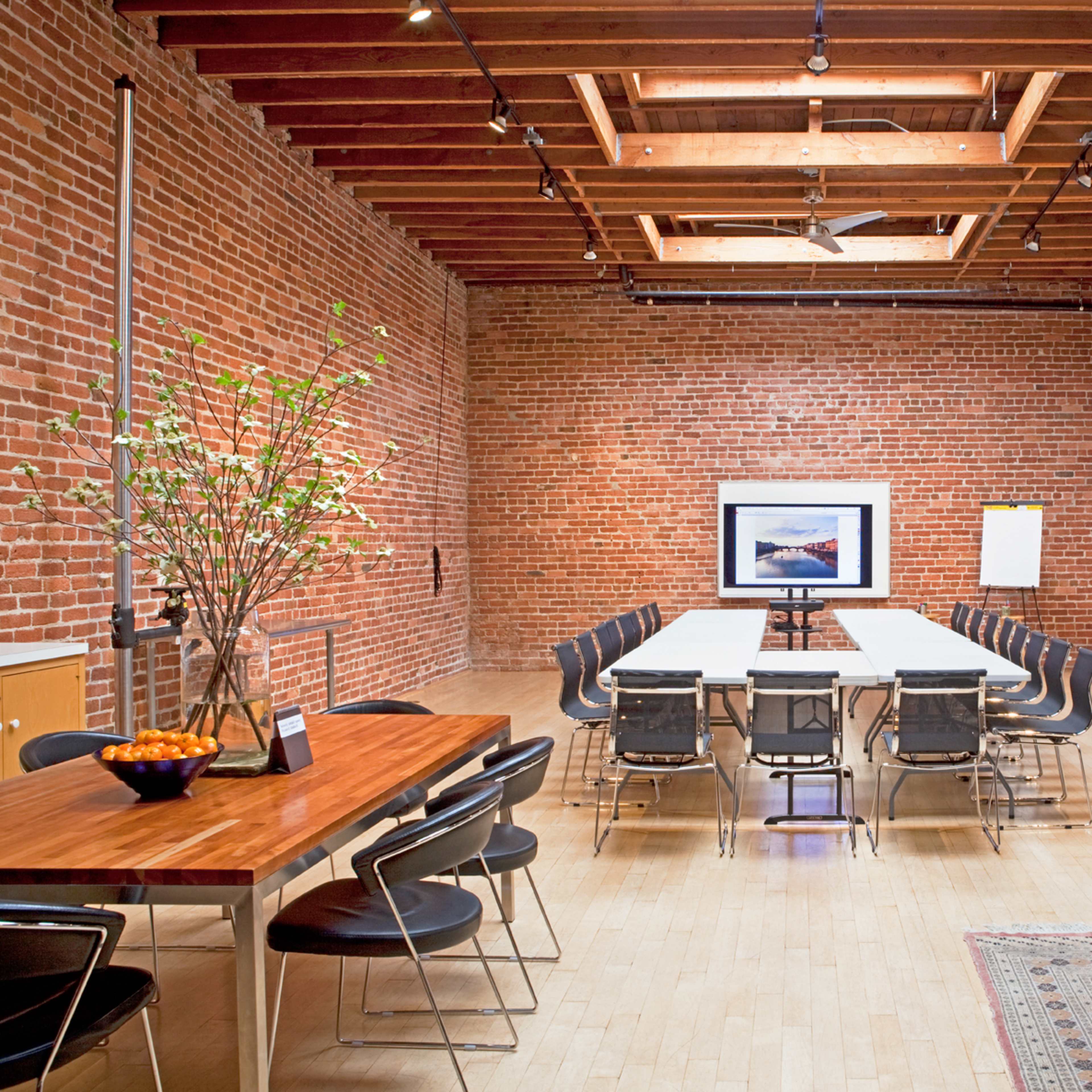 A spacious room with exposed brick walls, a wooden beam ceiling, a long central table surrounded by chairs, and various pieces of art displayed on the walls.