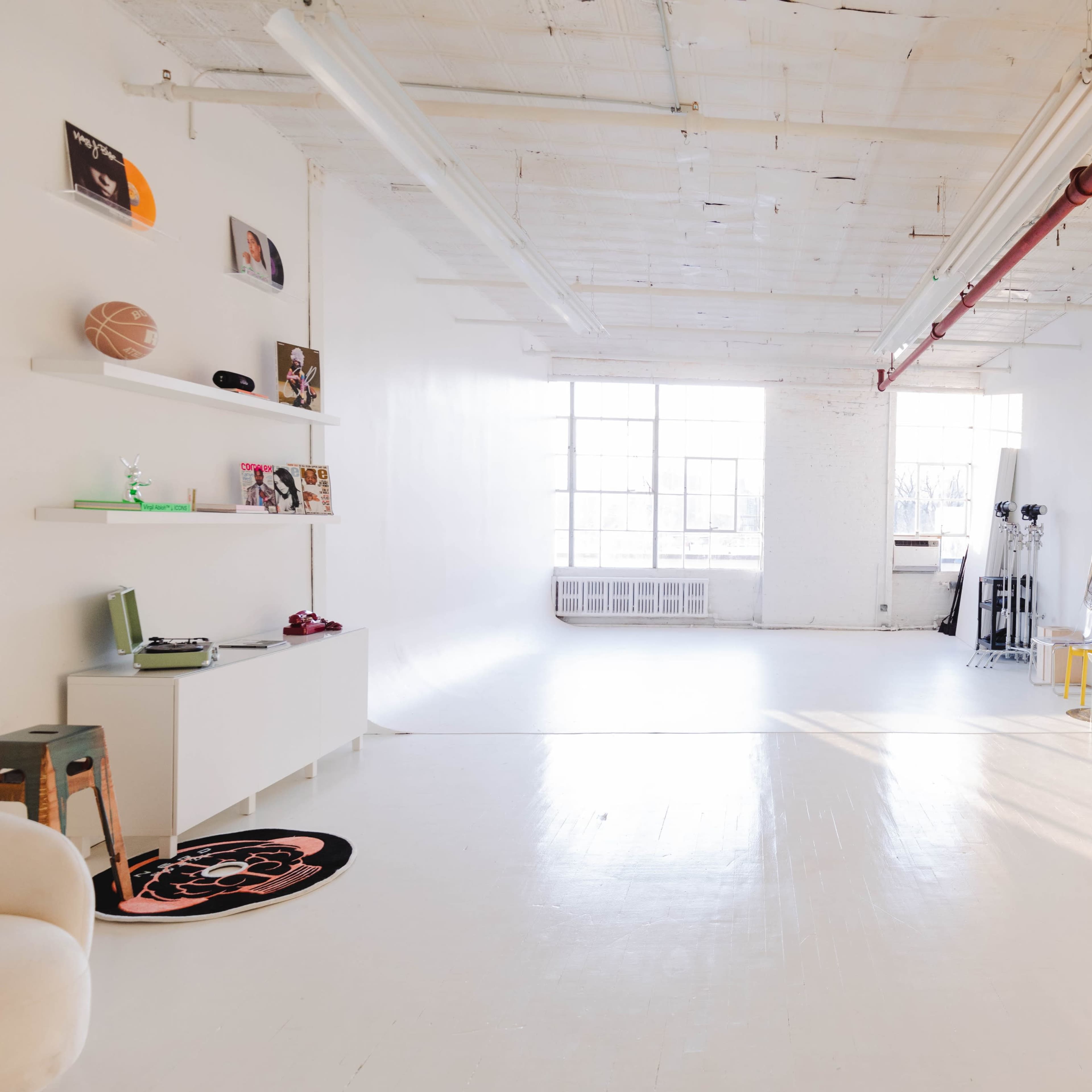 A bright, minimalist studio space with large windows, a white floor, and a few decorative shelves displaying items on the walls.